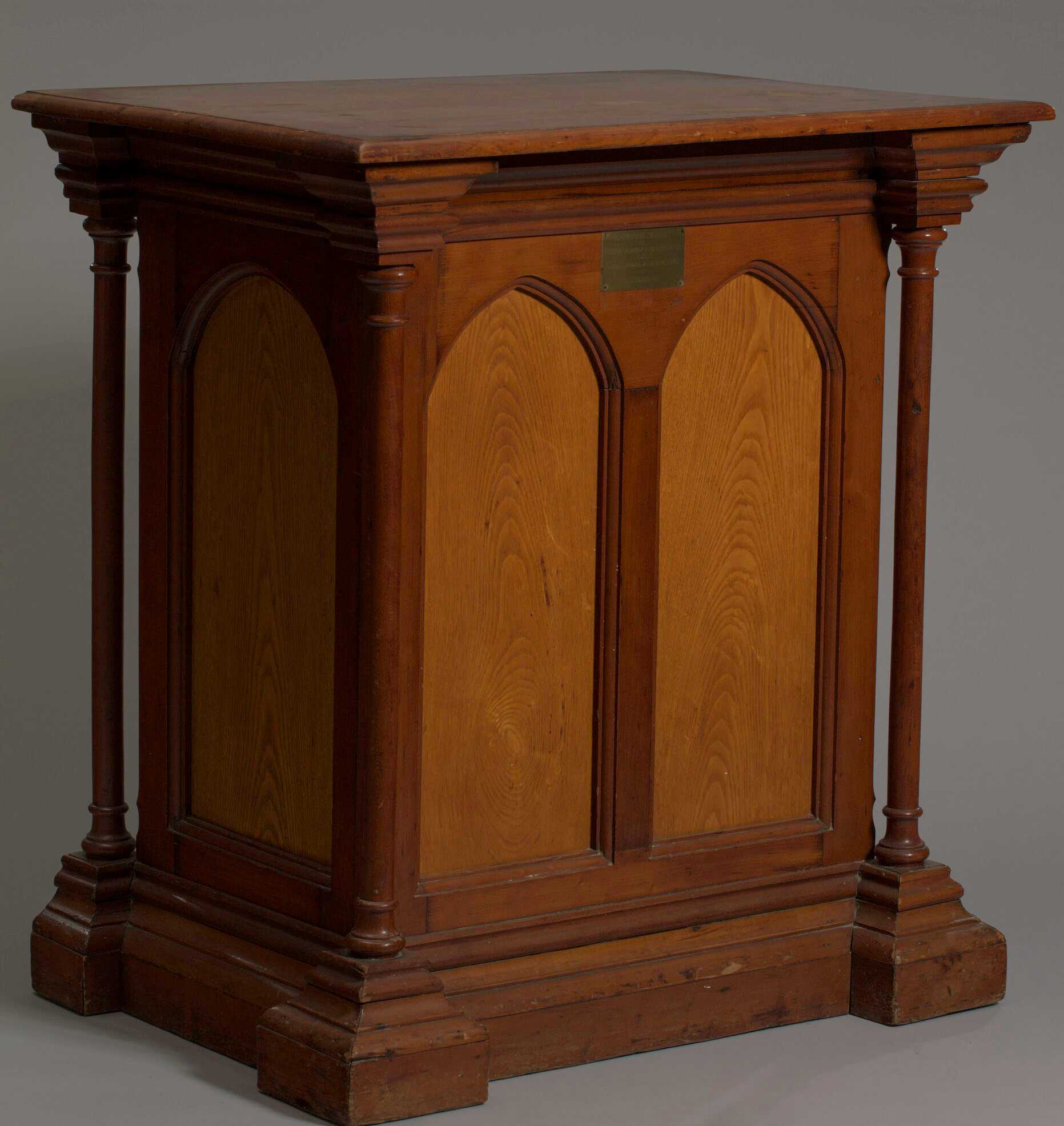 A pulpit from the Metropolitan AME Church, consisting of a hollow, three-sided structure composed of multiple pieces of wood that show different techniques, ages, and varnishes. The front of the pulpit features two arches separated by a piece of wood and framed by thin columns on either side. The wood is smooth and varnished with a light resin through which the wood grain is still visible. The cornices at the top and bottom of the columns are similar in style, with those on the bottom heavier due to extra supporting wood. There are small juts of wood in the interior suggesting a shelf may have been in place inside the pulpit for holding objects. A small metal placard screwed to the front of the pulpit notes “This podium was built for Metropolitan AME Church formerly Union Bethel AME Church by John Simms 1838.”