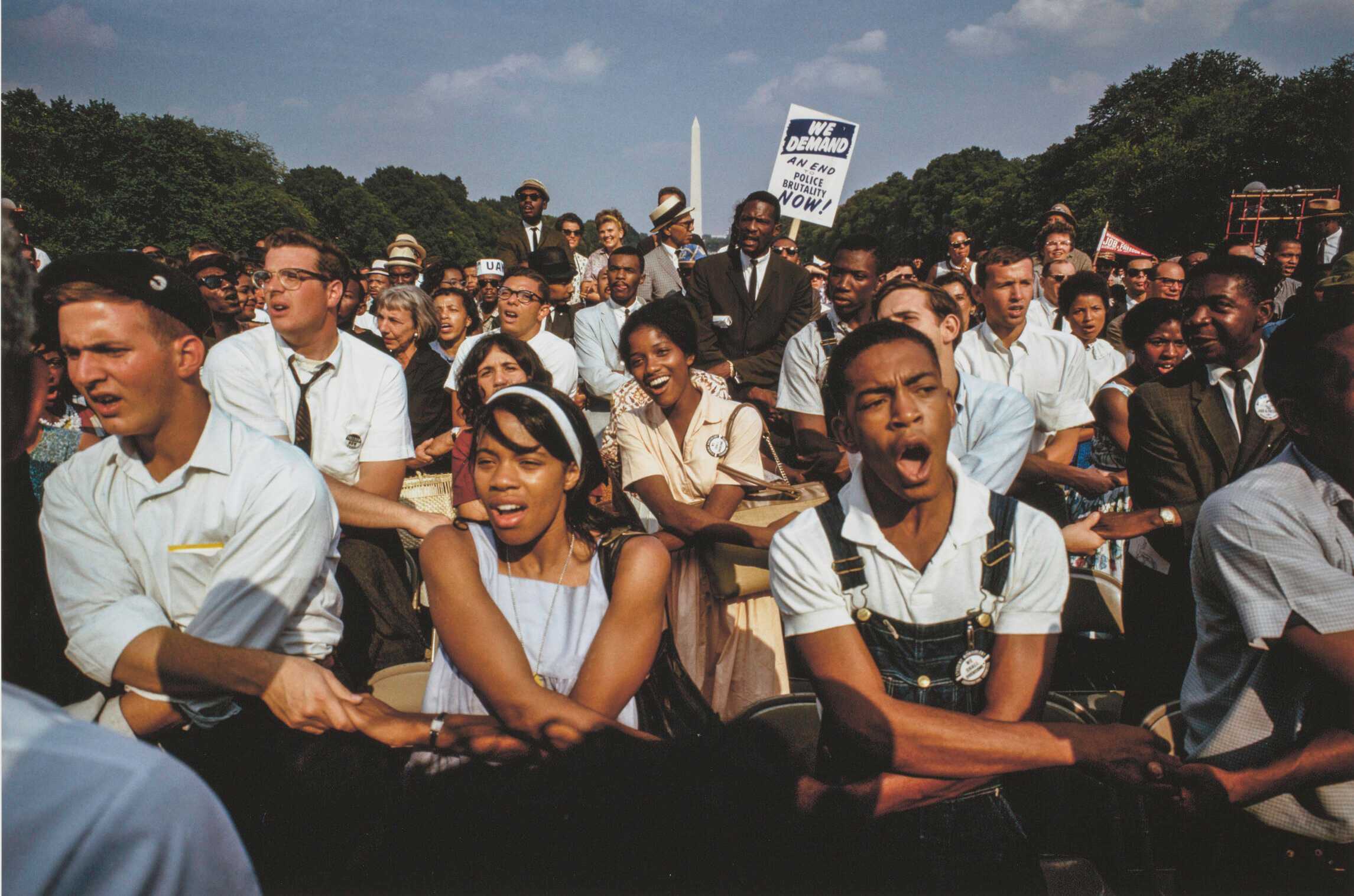 A color photograph by James P. Blair of the crowd of participants at the 1963 March on Washington. The image depicts a large crowd of different ages, genders and races gathered between the steps of the Lincoln Memorial and the Lincoln Memorial Reflecting Pool. The Washington Monument is visible in the far background. In the foreground of the image are rows of seated men and women with arms crossed in front of them and holding hands with the people on either side of them. Someone in the crowd holds up a placard that reads “WE DEMAND / AN END / TO / POLICE / BRUTALITY / NOW!” The print is signed by the photographer beneath the image at bottom right: [James P. Blair / 9 or 9]. Printed along the bottom edge is the caption and a copyright notice in black text.