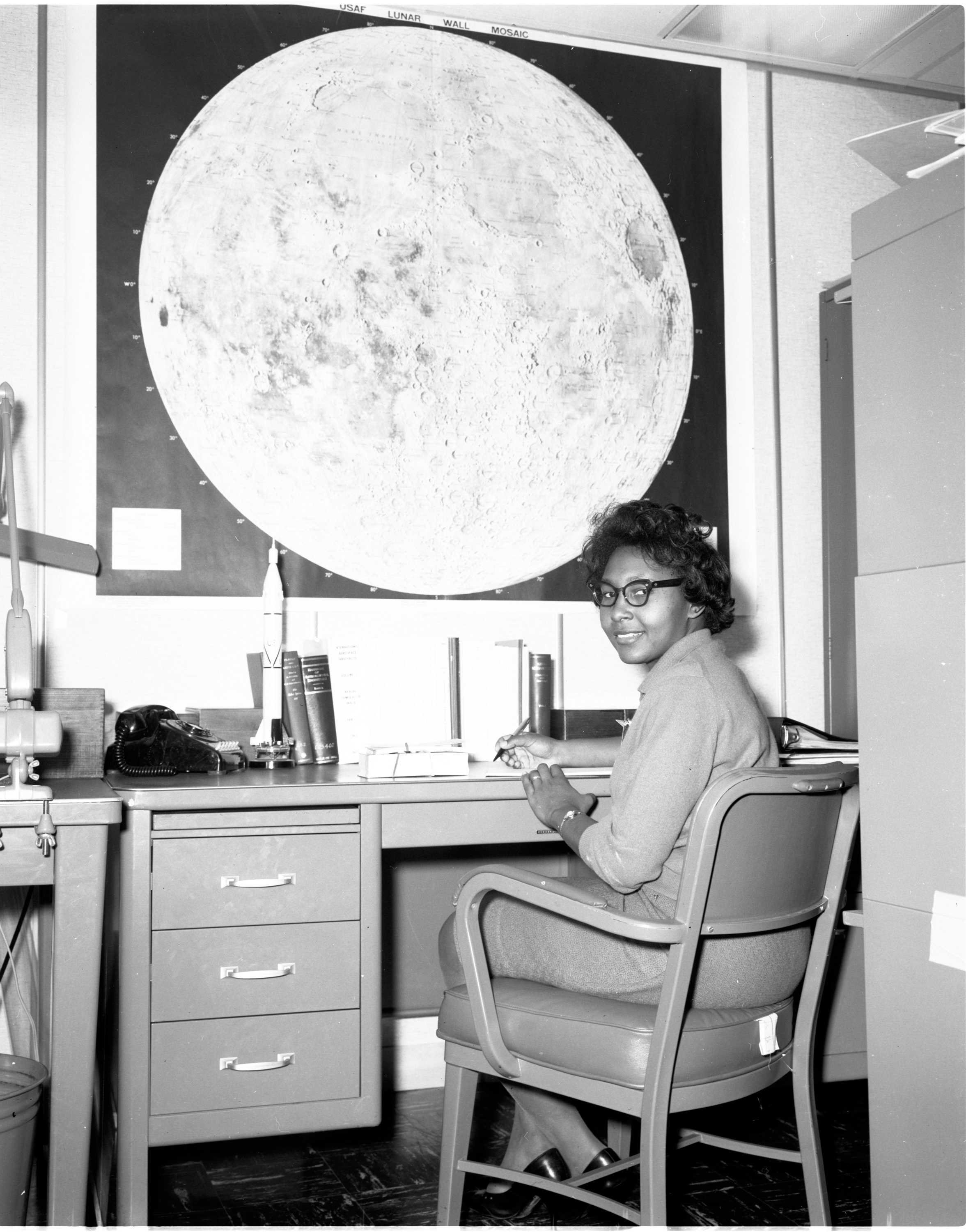 A black and white photograph of a Jeanette Scissum at a desk in front of a large poster of the moon.