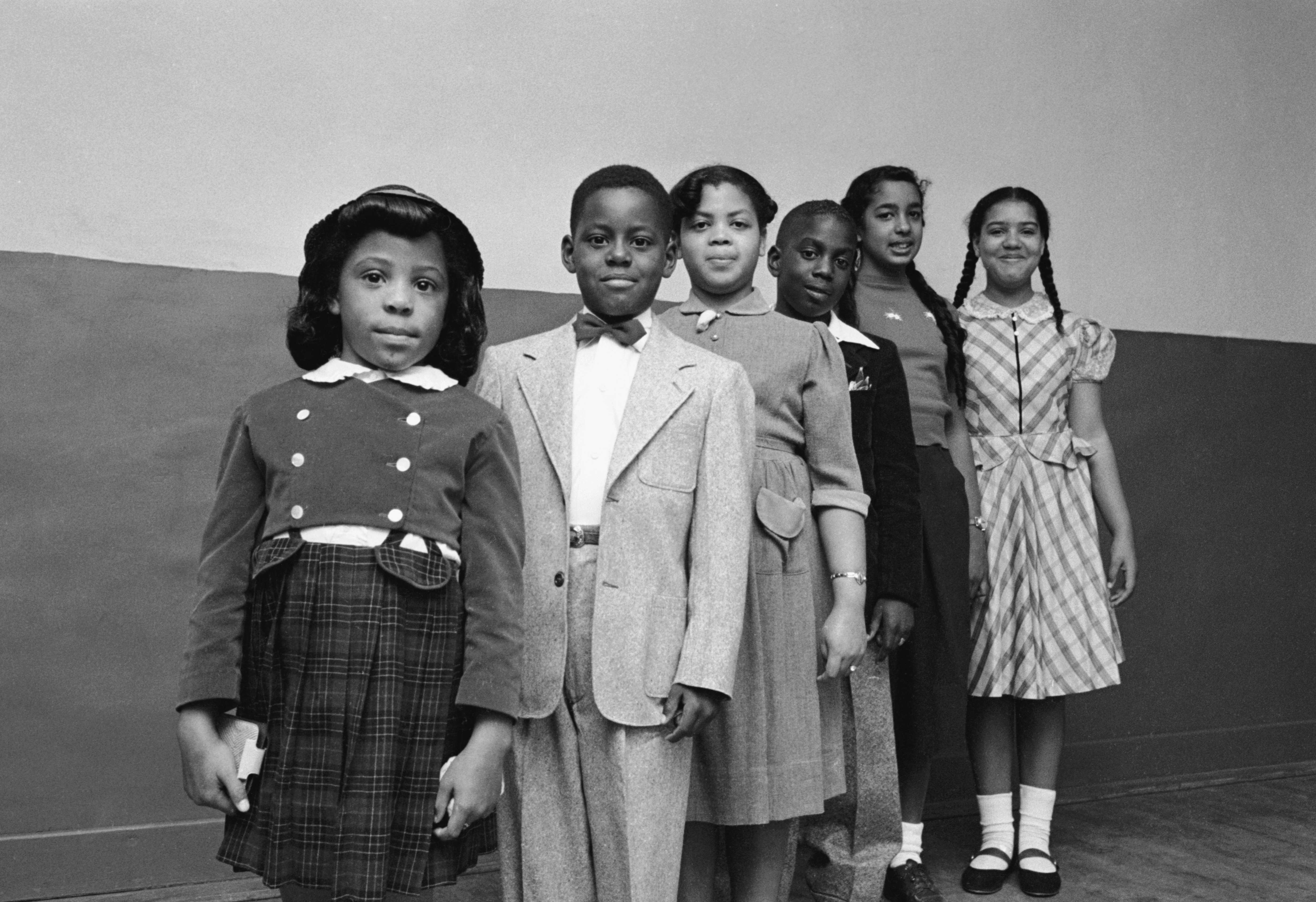 Black and white photgraph of 6 Black children standing in a line with hands by theri side.  There are 2 boys and 4 girls pictured.  The children are dressed up for school and look directly at the camera