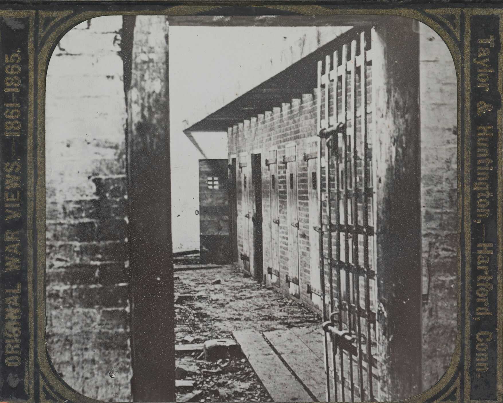 A glass lantern slide of a slave pen on the property of slave dealers Price, Birch & Company, where enslaved persons would be held until auction. The image depicts the view through the outer gate into interior of a slave pen. To the left, a building with a series of narrow doors, each with a small barred window. The printed slide casing reads [ORIGINAL WAR VIEWS 1861-1865.] along the left side and [Taylor & Huntington, - Hartford, Conn.] on the right.