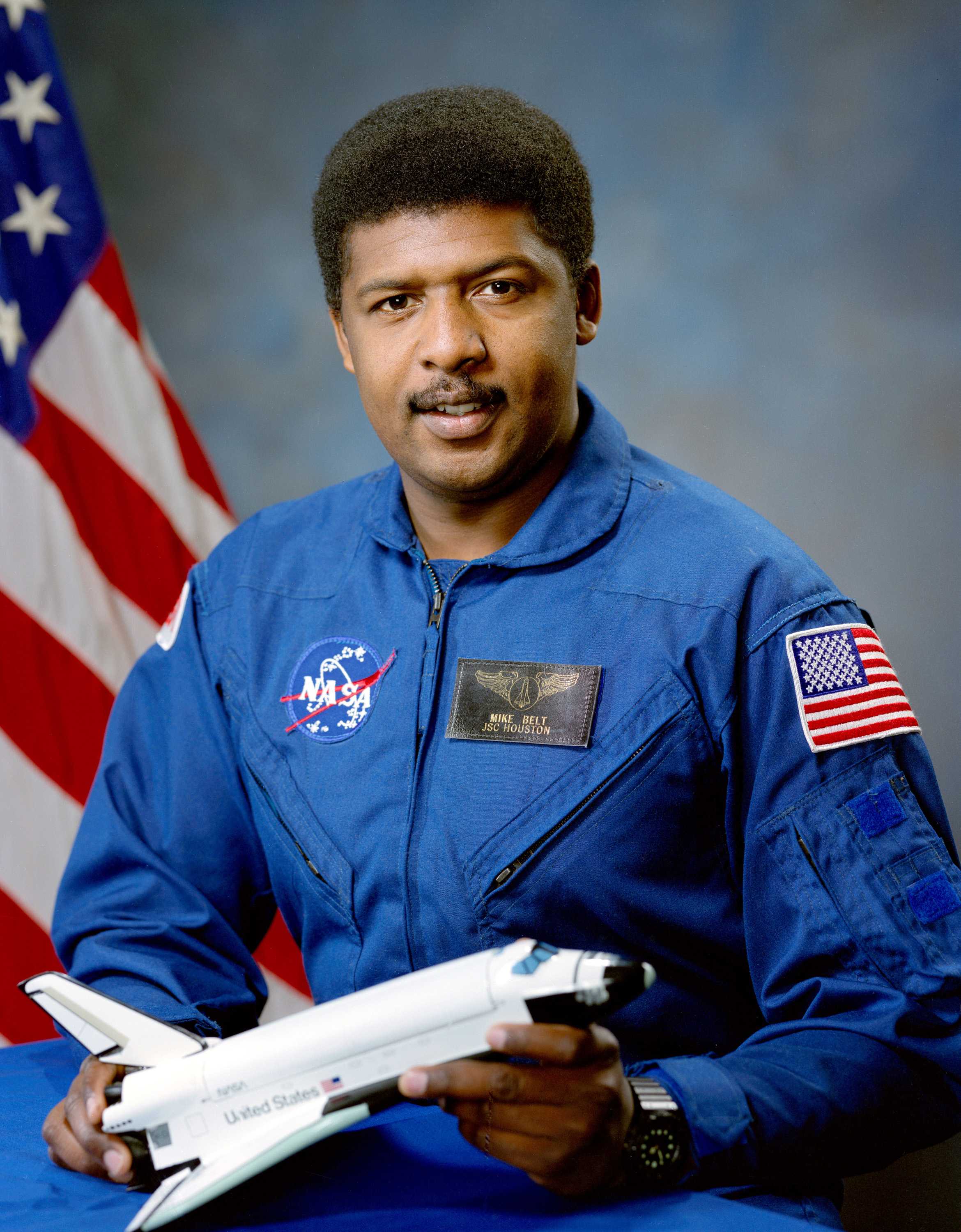 Michael Belt is wearing a blue NASA uniform with a model of a spaceship in his hands as he smiles for the portrait.