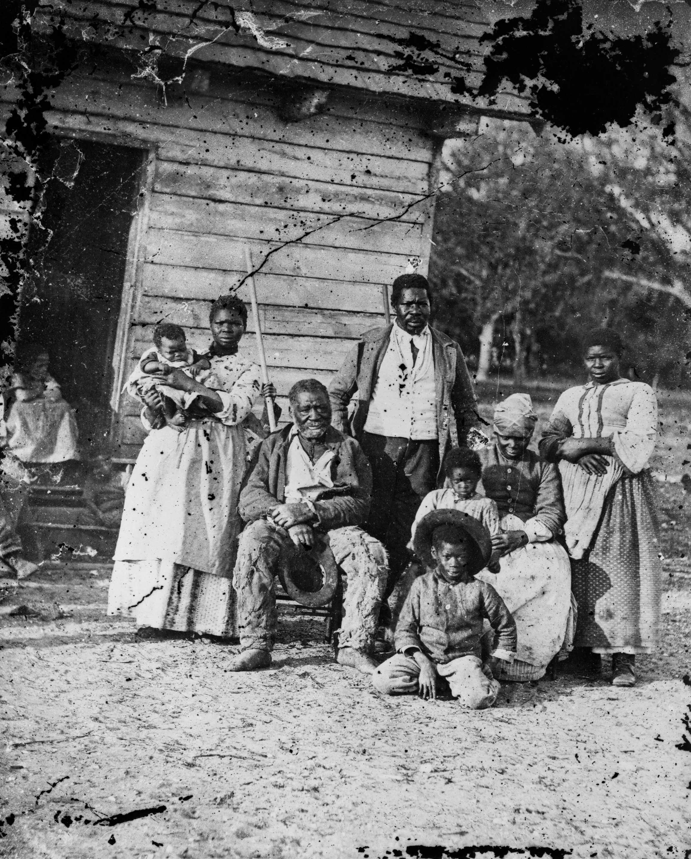 A black and white photo of a family of 4 adults and 3 children posed for a picture outside of a cabin.
