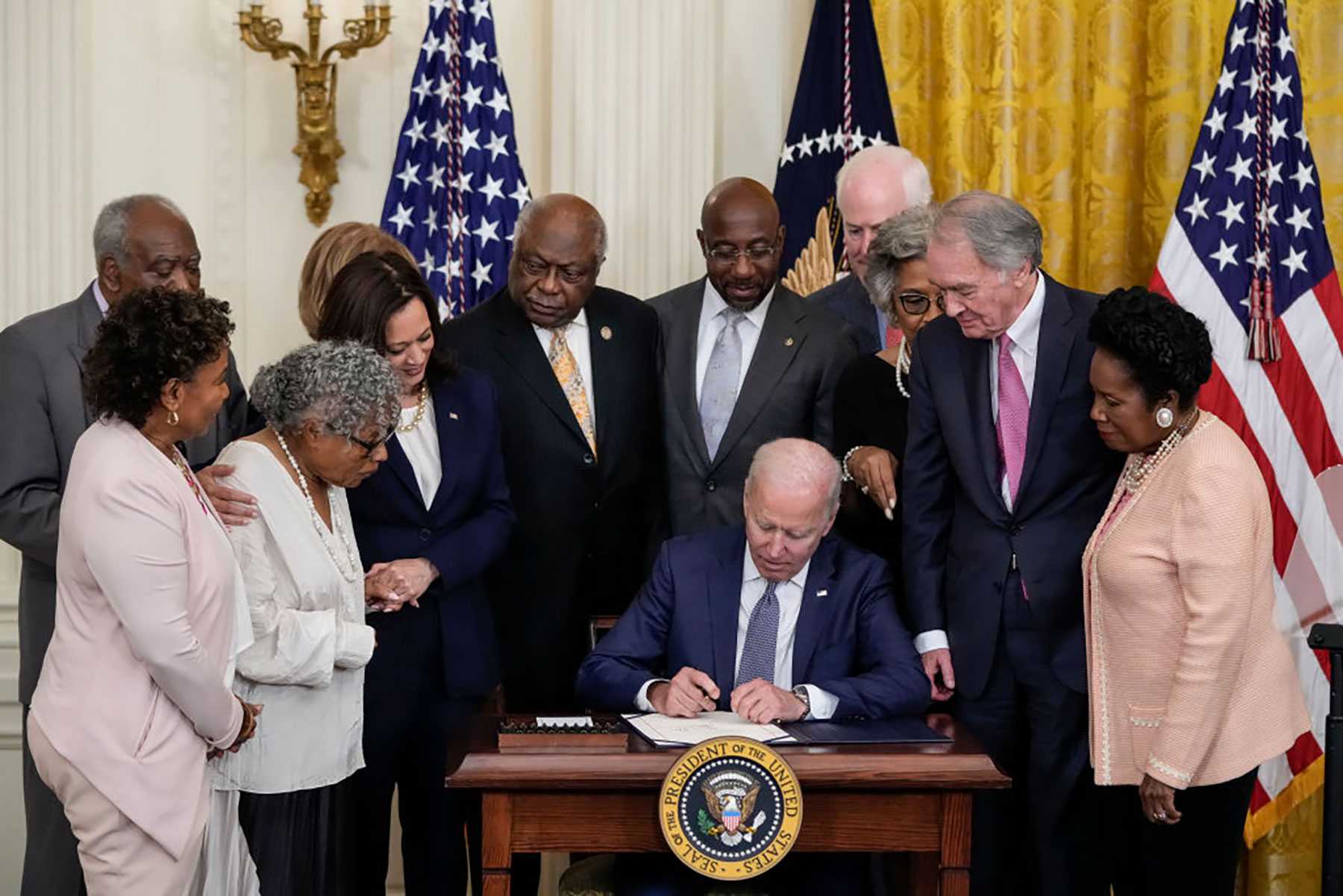 A group of people stands around a man at a desk signing a document, with flags in the background.