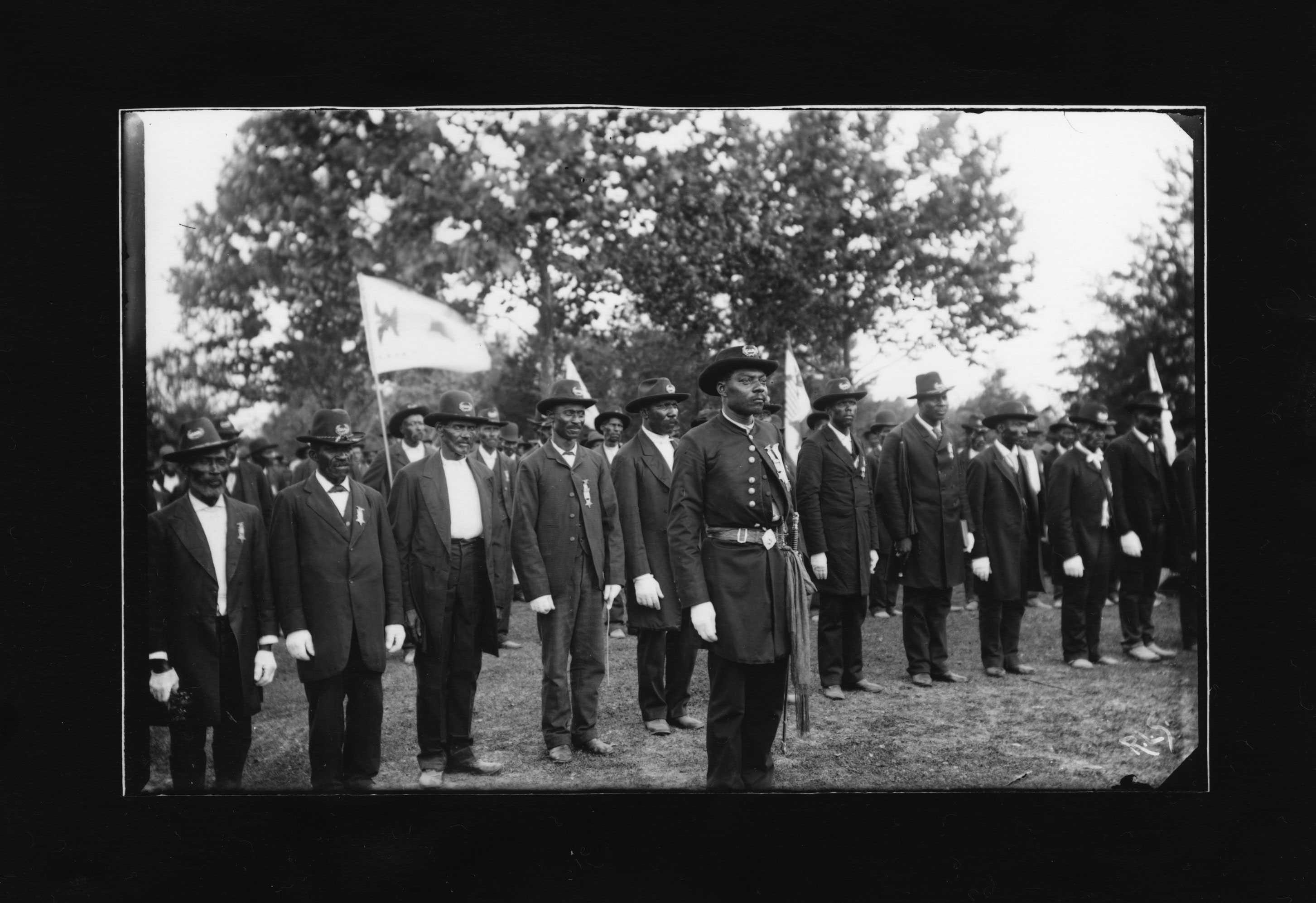 A black and white photo of 2 rows of African American soldiers lined up next to each other. Some hold guns and flags.