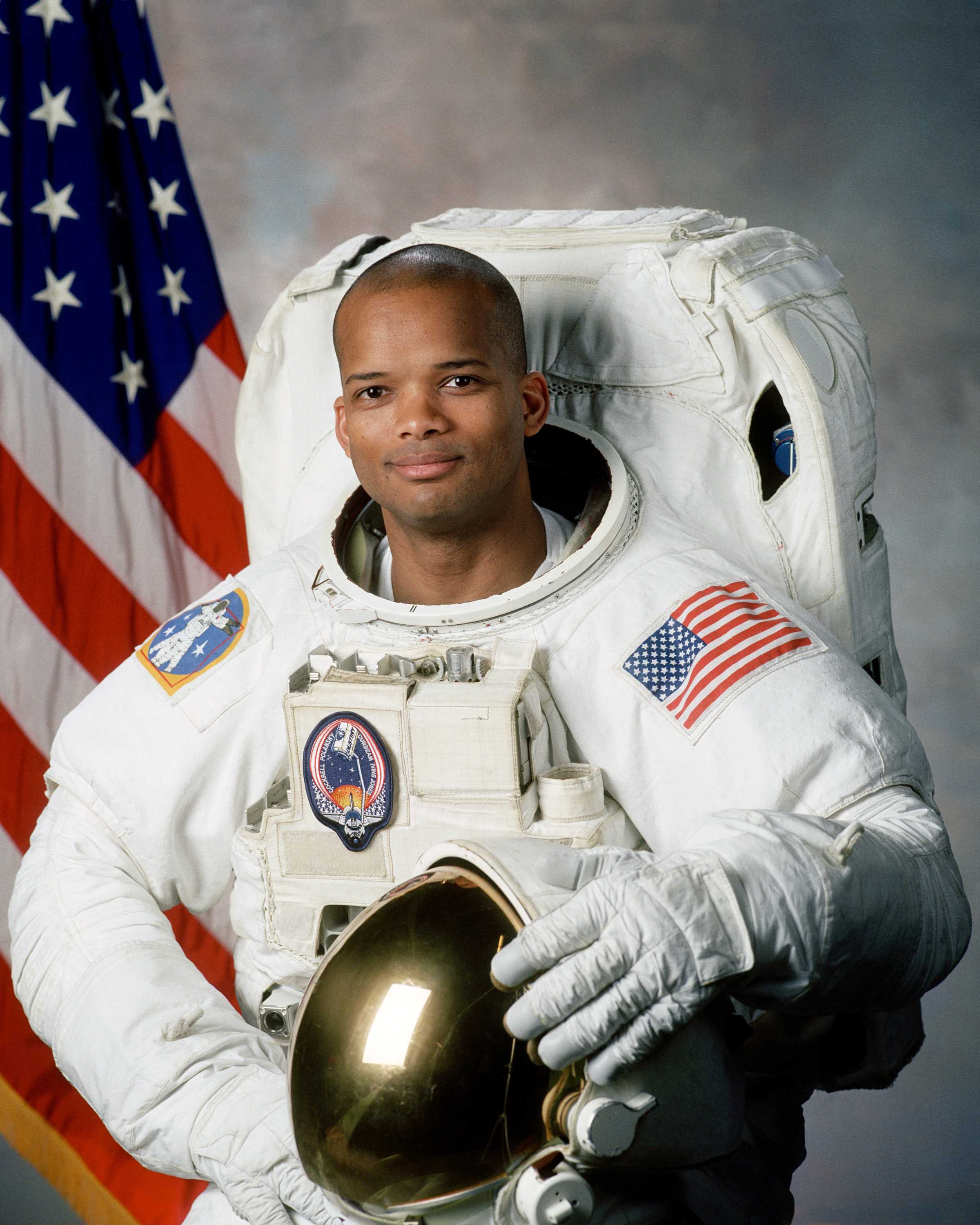 Robert Curbeam poses for a portrait in a white NASA space suit while holding a helmet.