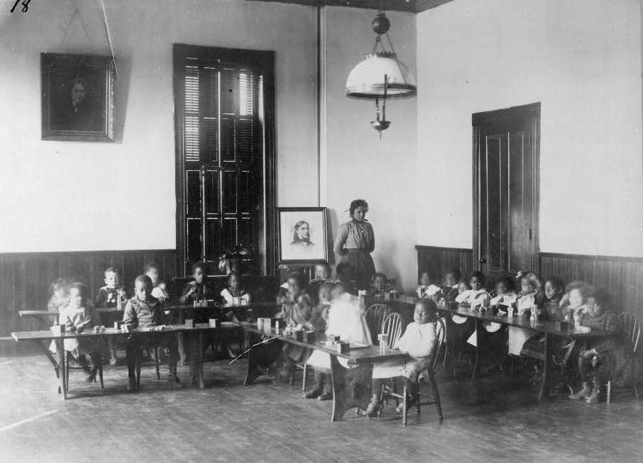 Black and white photograph of classroom with kindergarten students seated at 2 L shaped long desks.  A woman (teacher) stands in the corner behind them next to a framed portrait of unidentified man.