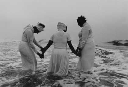 A black-and-white photo of three women in white dresses standing in ocean waves during a memorial ritual.