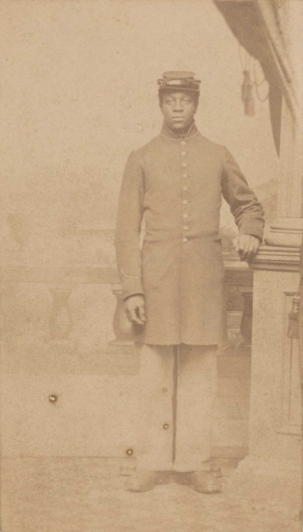 A reddish-brown photograph of an unidentified Union soldier posed in a photographer’s studio. The man is standing slightly off-center and is wearing a nine-button frock coat with a high collar. The coat has contrasting piping around the collar and sleeves. He is also wearing straight trousers, heavy boots, and a slouched kepi hat with a leather band. He is standing, looking straight at the camera, in front of a colonnade railing with fluted rock supports. Though it is too faded to see, there is presumably an idyllic city or landscape behind him. His left hand is propped on a low riser next to a column topped with a hanging flag and tassels. The back of the photograph has a studio mark at center that reads “Photographed by/ CUSHING,/Woodstock, VT.” Also printed on the back at the bottom of the card is “Negatives Preserved.”