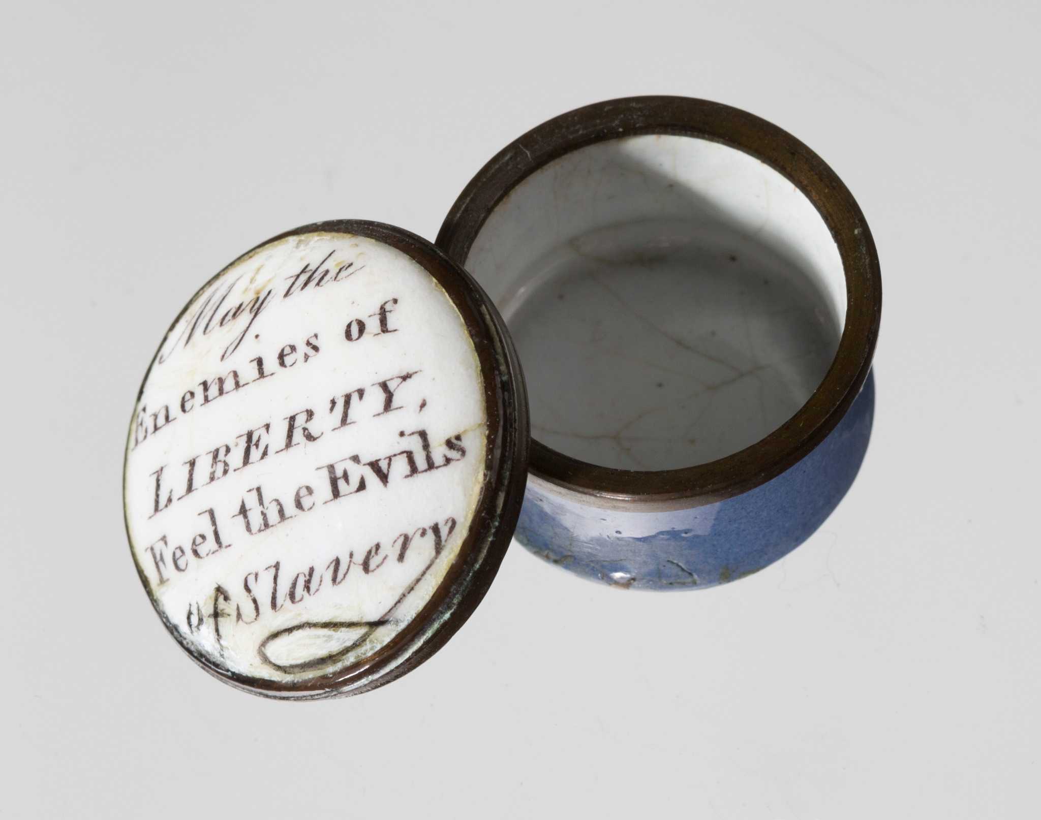 A round, blue, white and black enamel container with a removable lid. The lid of the container is white. Written in black script on the lid of the container is "May the Enemies of LIBERTY, Feel the Evils of Slavery." The outside of the container is light blue, with a black rim. The inside is white.