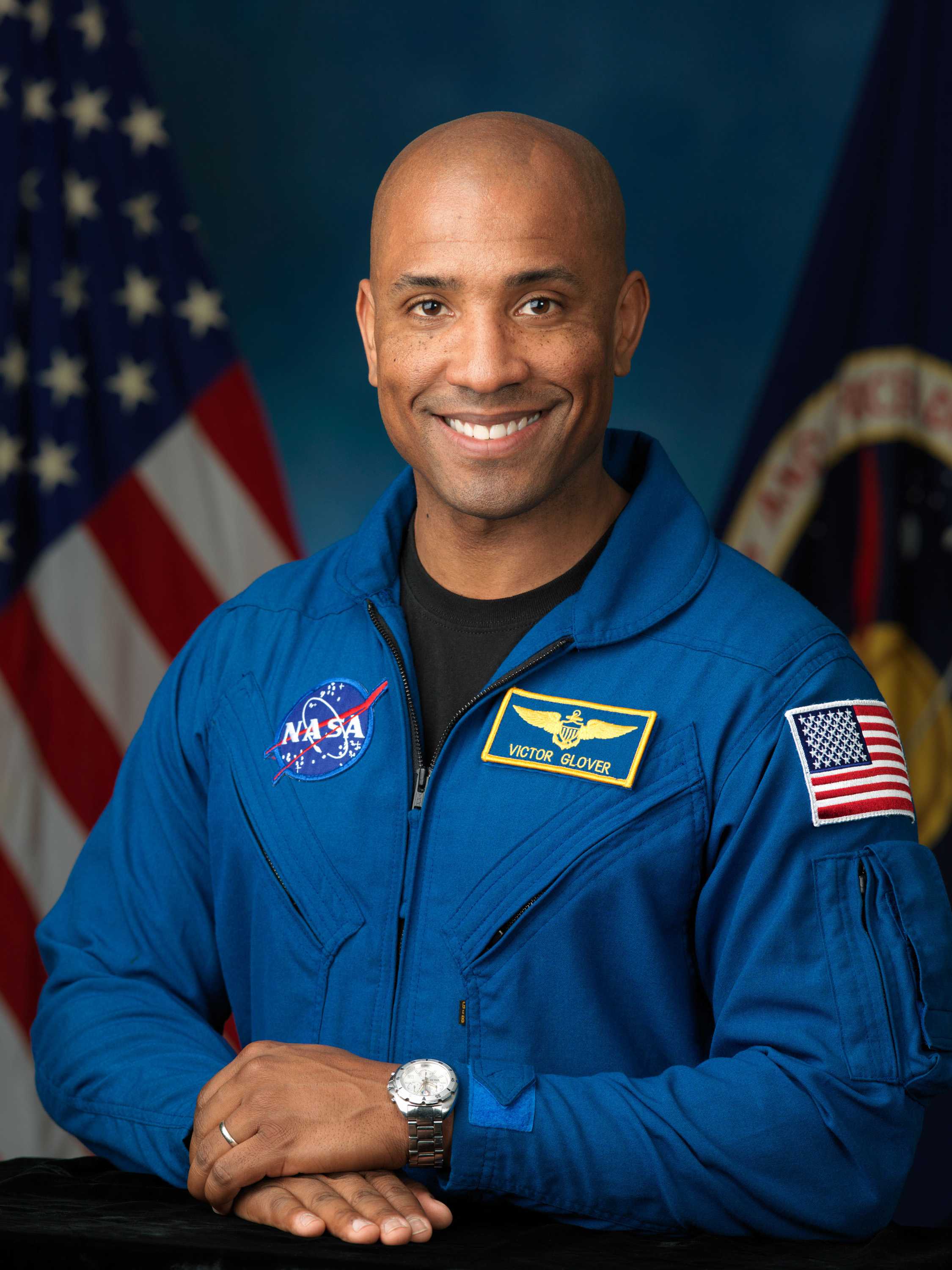 Victor Glover is smiling while he poses for a portrait in a blue NASA suit with his hands crossed.