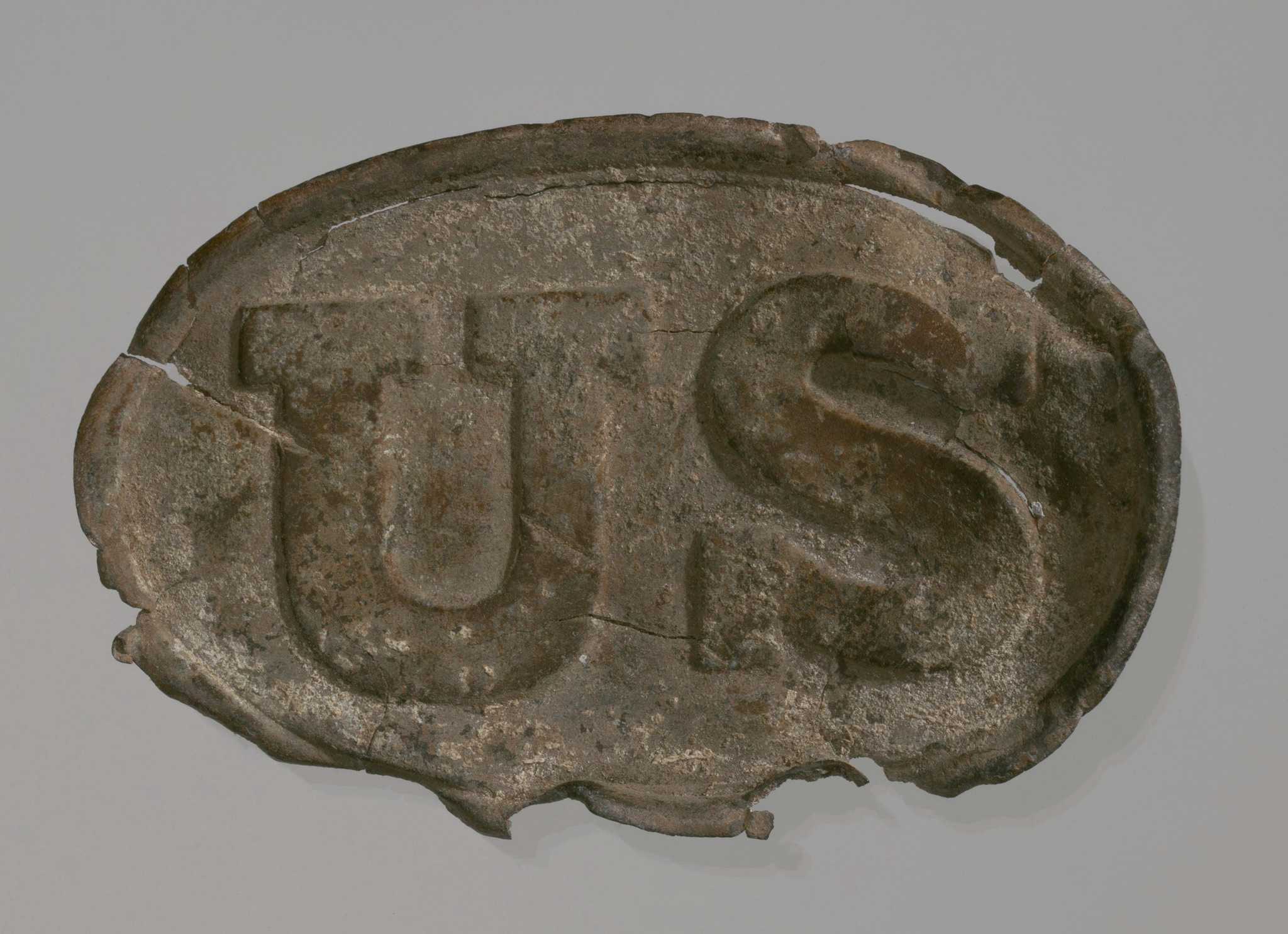 A metal Union belt buckle. The metal buckle is oval shaped with the raised lettering on the front that reads [US]. There are three hooks on the back. Any backmarks that may have existed were worn away by the elements.