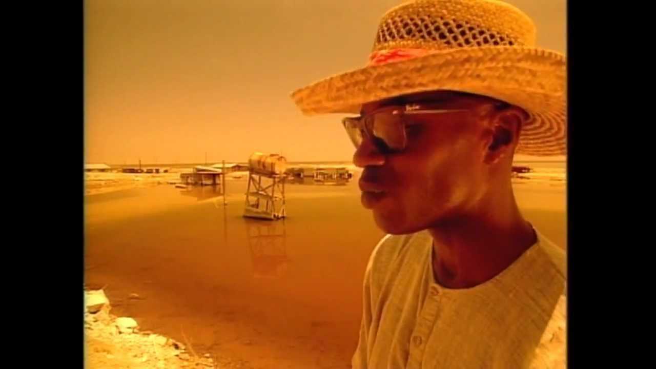 With an orange overlay, John Akomfrah is on a beach in a straw hat, sunglasses, and white shirt.