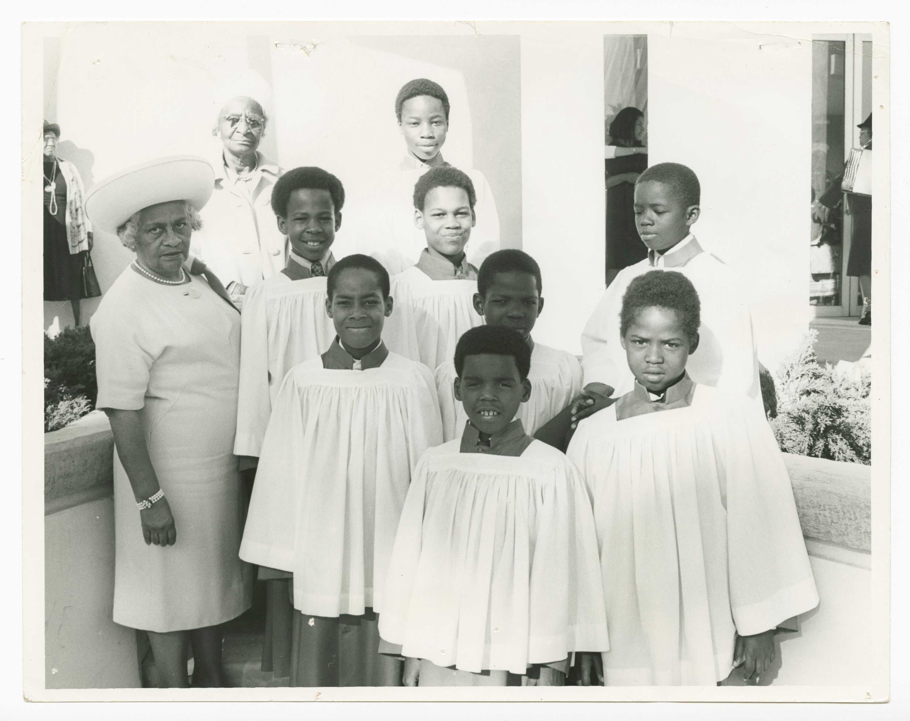 Black and white photograph of a group of young boys dressed in altar clothes. The group includes eight (8) boys in several different rows. The boys wear white robes and dark skirts and dark collars over their clothing. On the left side of the image are two (2) older women wearing white looking at the camera, one in the foreground and one in the background. the group stands outside with the church interior seen in the upper right quadrant. On the reverse in the lower right corner on the bottom in pen [ALTAR BOYS].