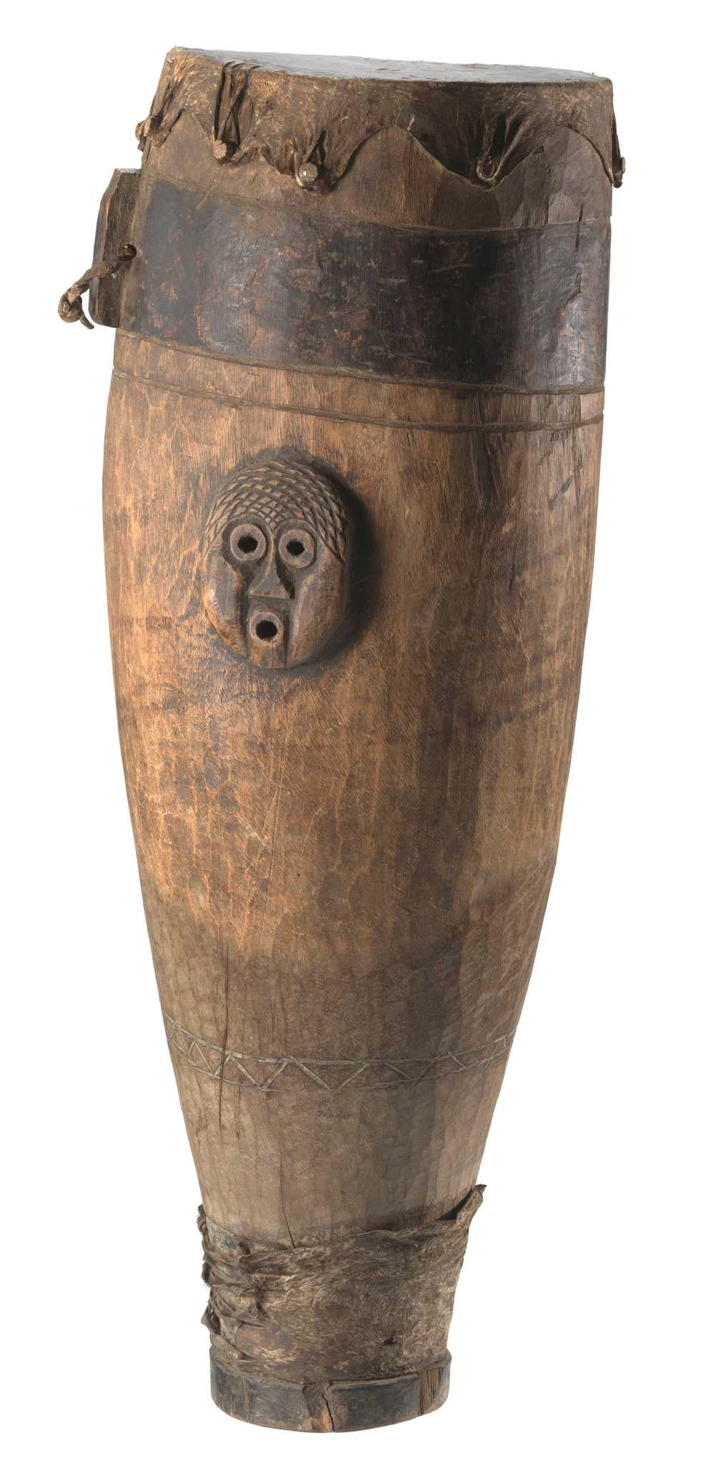 A wooden drum covered with stretched leather hide that has been attached to drum body with nine wooden pegs. Two carved, anthropomorphized face motifs are on opposite sides of the upper half of the drum body. The edges of face motifs have been sealed with a darker adhesive material. There are three bands running the circumference of the drum. The top band closest to the drum head, is stained darker than the rest of the drum body. There is a carved piece that extends from the darker band. It has a round drill hole  that had been threaded through with a leather thong. This piece has been stained the same color as the bank. The middle band is directly below the top band and above the face motifs. It is the same color as the rest of the body. The bottom band is a carved triangular pattern between two parallel lines. The bottom band is located near the base of the drum, above a piece of hide wrapped and tied around the very bottom of the drum body. There is a nail visible in drum body interior.