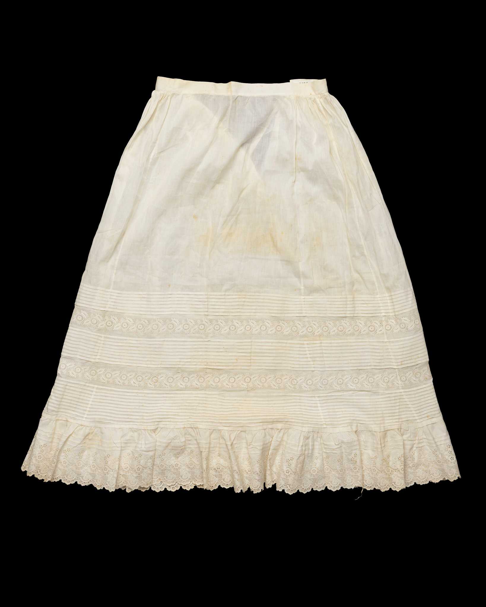 A handsewn tucked petticoat with embroidered detailing. The white petticoat has a tie closure at the waist. The lower half of the petticoat has multiple tiers of tucked detailing alternating with rows of embroidered flora. Along the hem is more elaborate embroidery with a scalloped edge. The fabric is discolored with small areas of loss. There is a handwritten label sewn in the interior waistband.