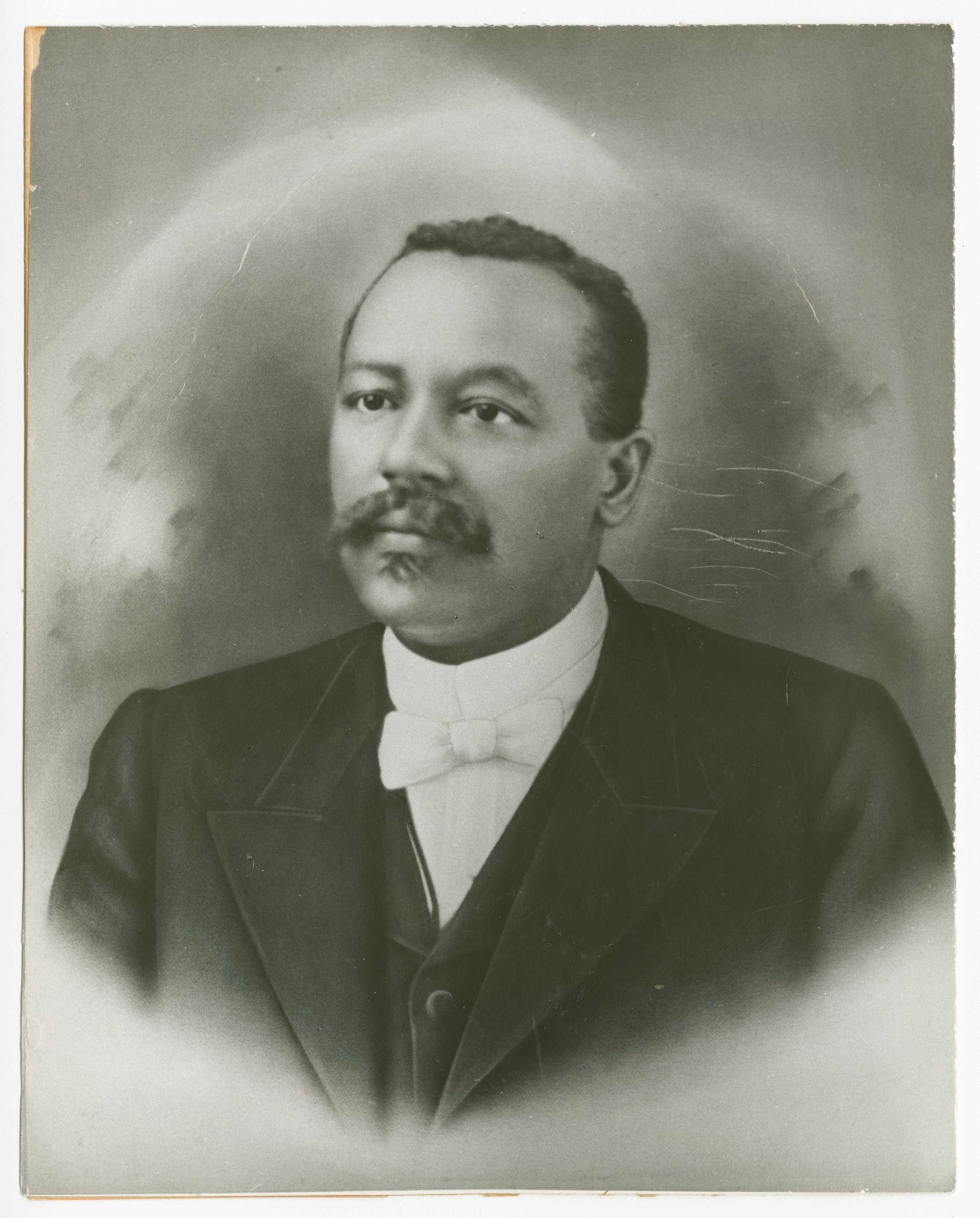 Black and white portrait image of a man wearing a dark jacket, dark vest, white shirt, and white bow-tie. He has a mustache and goatee and looks the left of the camera. The image is hazy at the corners and along the bottom. The background is darker near the man's head. The image shows the man from the chest up in three-quarter view. The photo is adhered via glue to a piece of cardstock the same size as the photo.