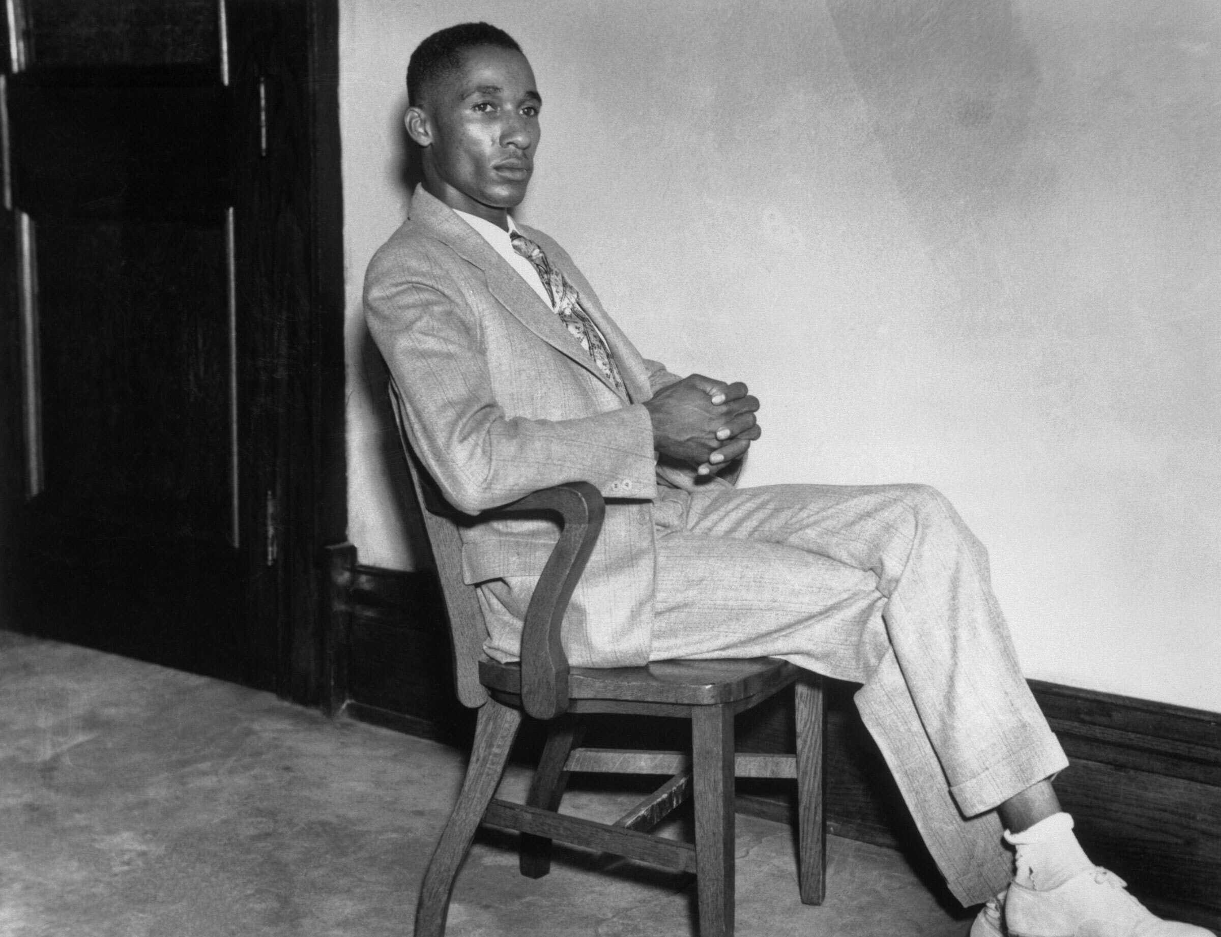 Black and white photograph of Black man seated in a wooden chair with crossed legs and hands folded in lap.  He is wearing a light colored suit and patterned neck tie.