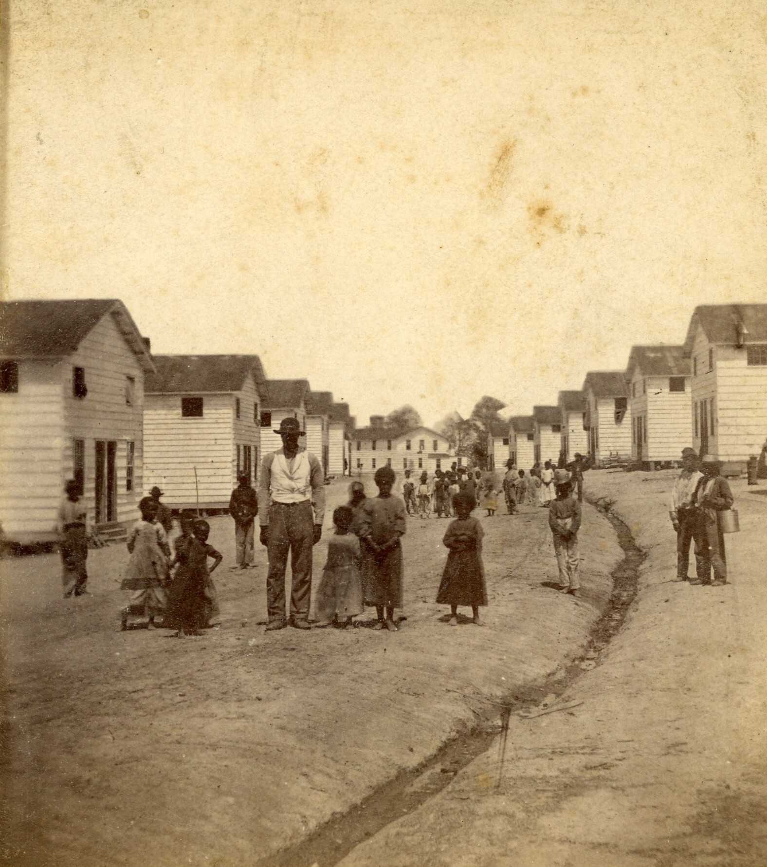 Historic photograph showing a dirt road lined with rows of identical white wooden houses. Groups of people, including adults and children, stand and walk along the road.