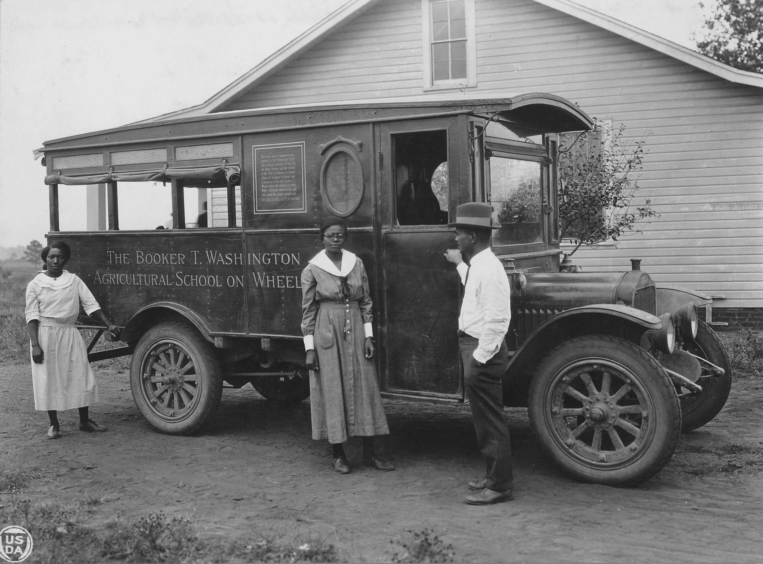 Photograph of three people near a bus labeled "The Booker T. Washington Agricultural School on Wheels."