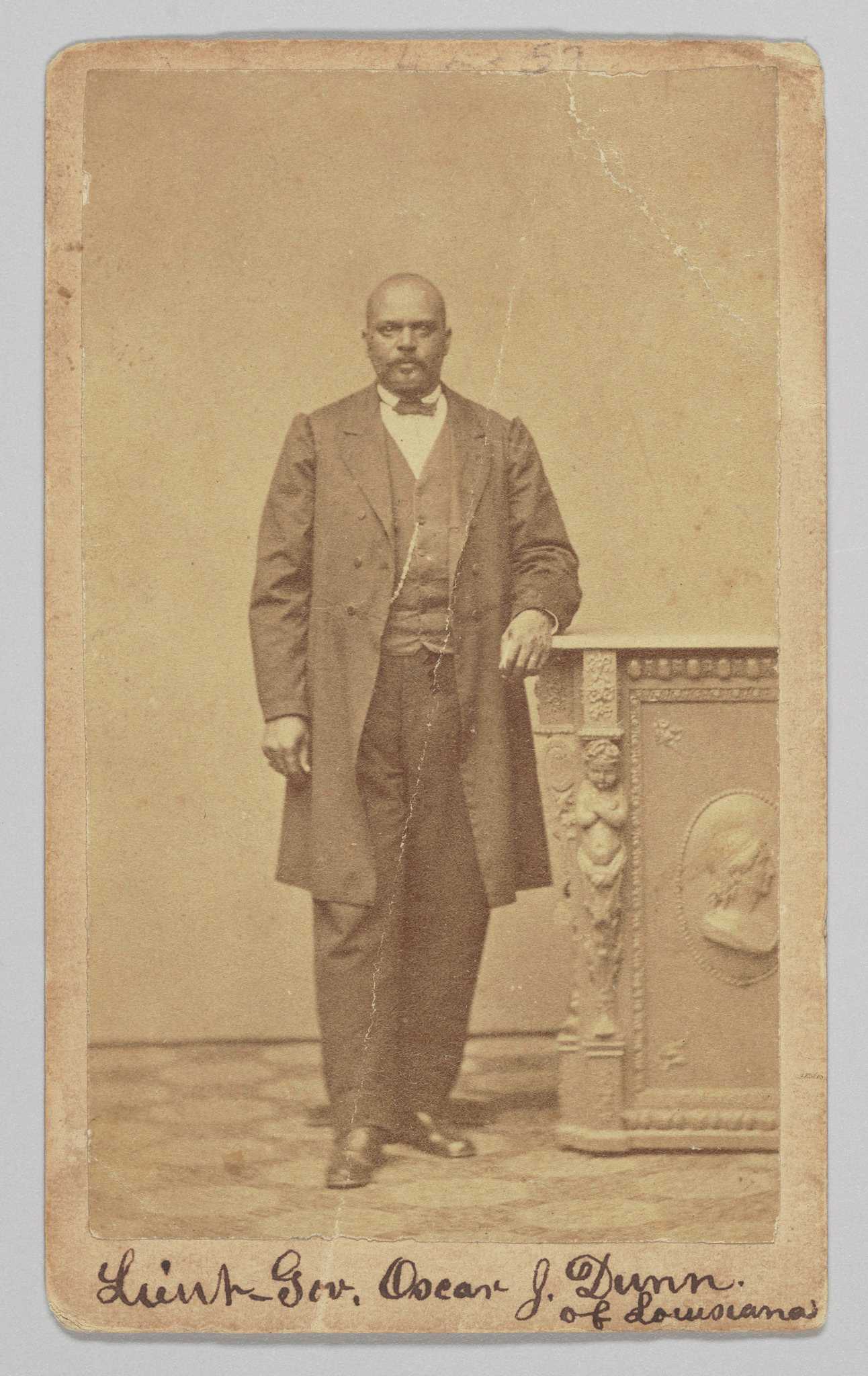 An albumen print carte-de-visite portrait of Lt. Governor Oscar J. Dunn. He is photographed standing with his right foot slightly forward than his left and has his left arm resting on a pedestal. He is wearing a dark colored suit, vest and a bowtie. A watch chain is visible on the right side of his vest. He is looking directly at the camera. The photograph is inscribed on at the top and bottom of the front. At the bottom, handwritten in black ink, is: [Lieut Gov. Oscar J. Dunn / of Louisiana].
