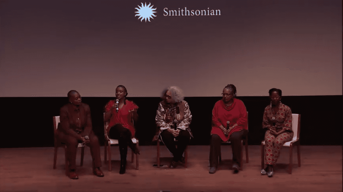Five black women speaking on a panel on the stage in front of a large screen.