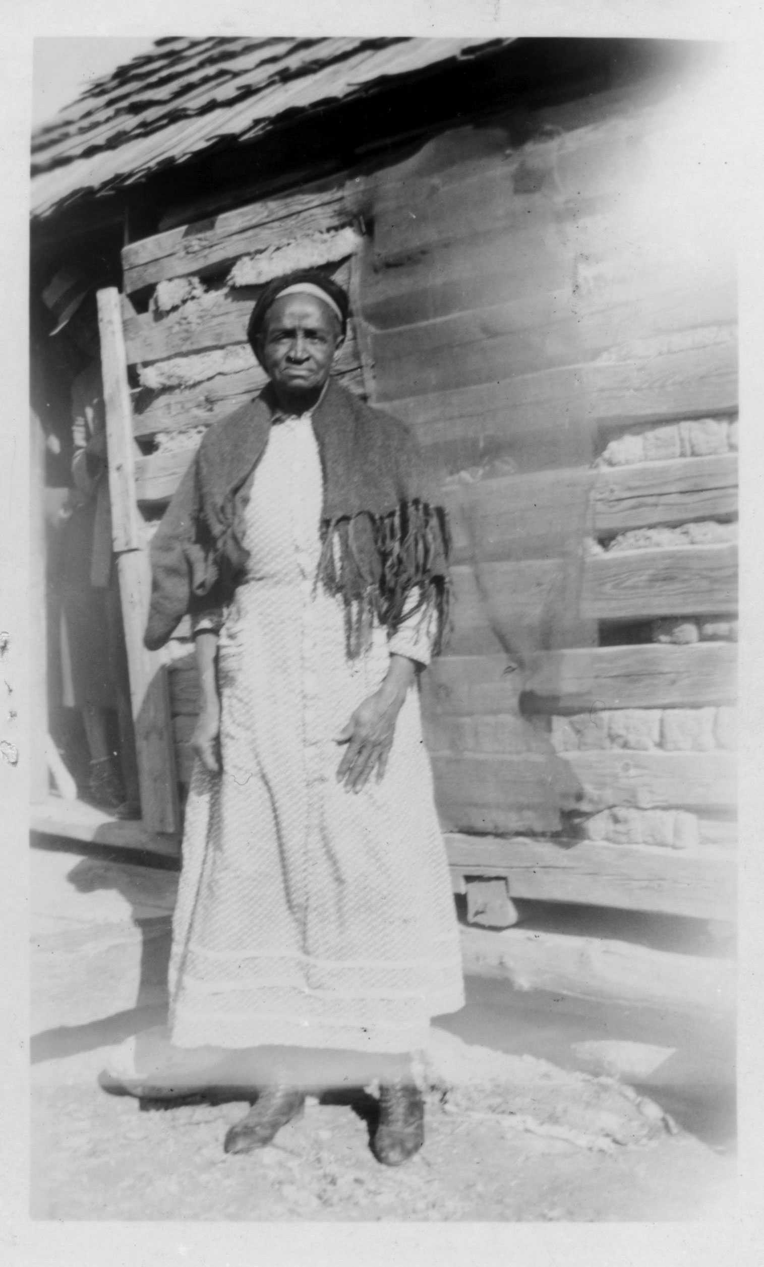 An overexposed black and white photograph of Lucy Thomas standing in front of a log cabin.
