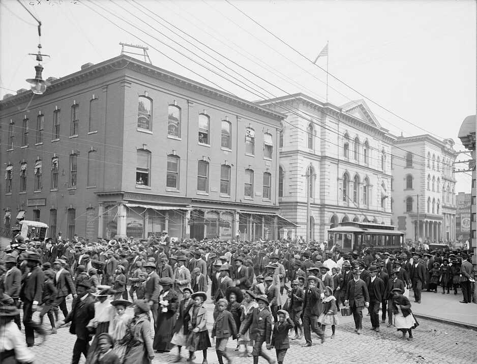 A black and white photograph of Emancipation Day festivities. A crowd of people walking in the streets downtown.