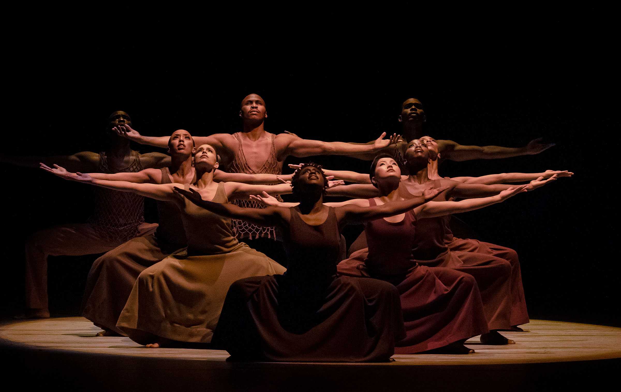 Dancers in a group pose on stage with outstretched arms.