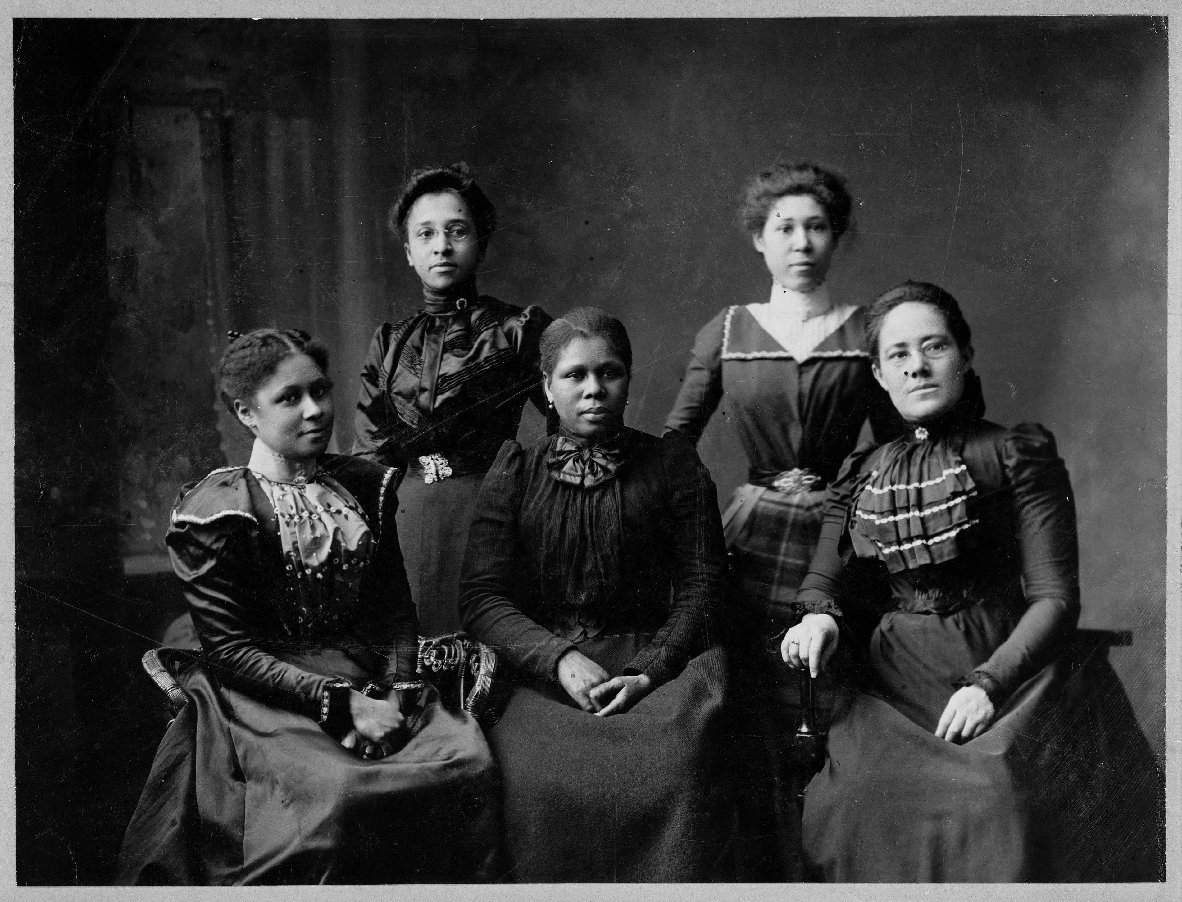 A black and white portrait of 4 Black women in a studio. Each is wearing a dark dress with their hair pinned back.