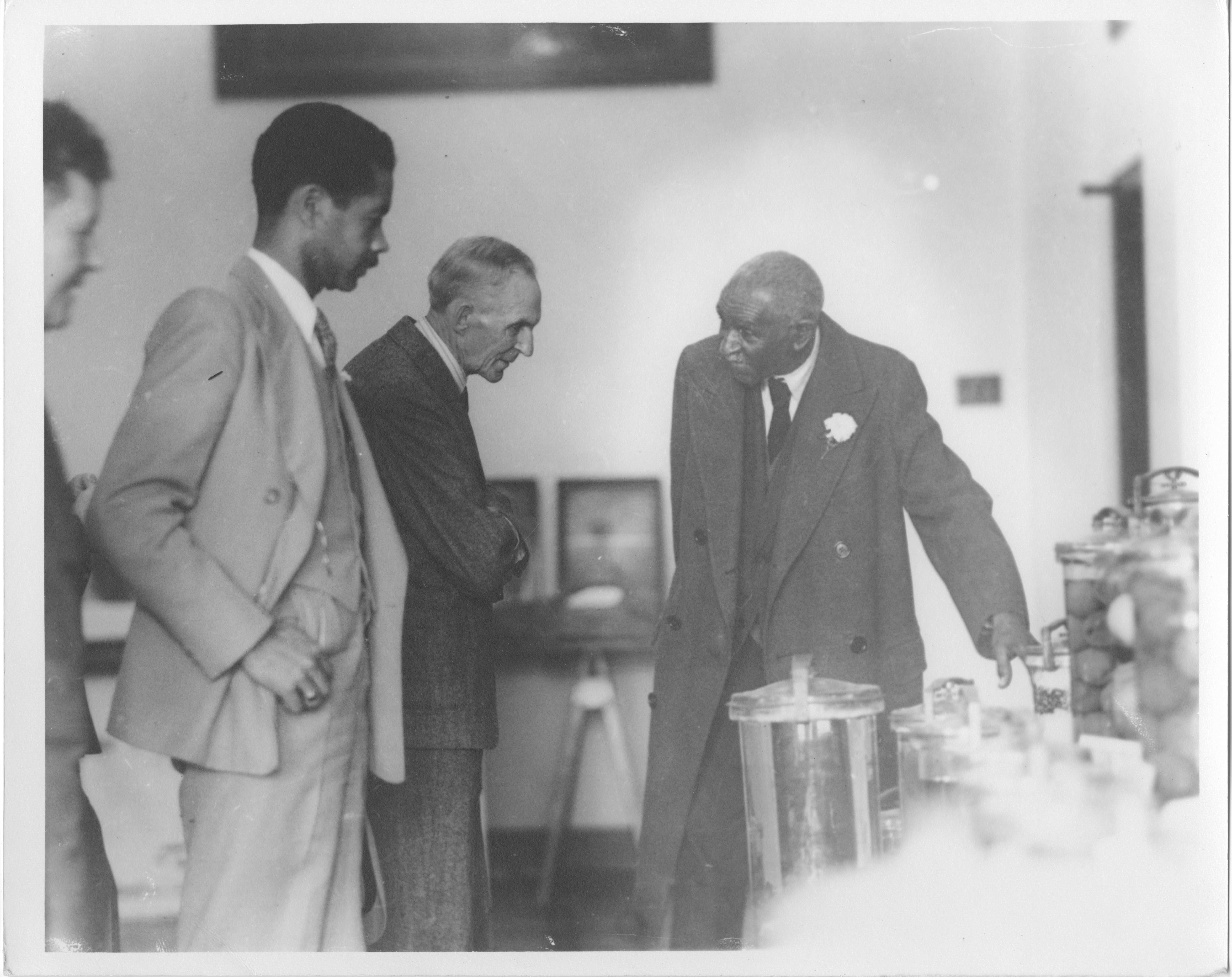 Black and white photograph of George Washington Carver showing Henry Ford his laboratory.  There are two unidentified men looking on.