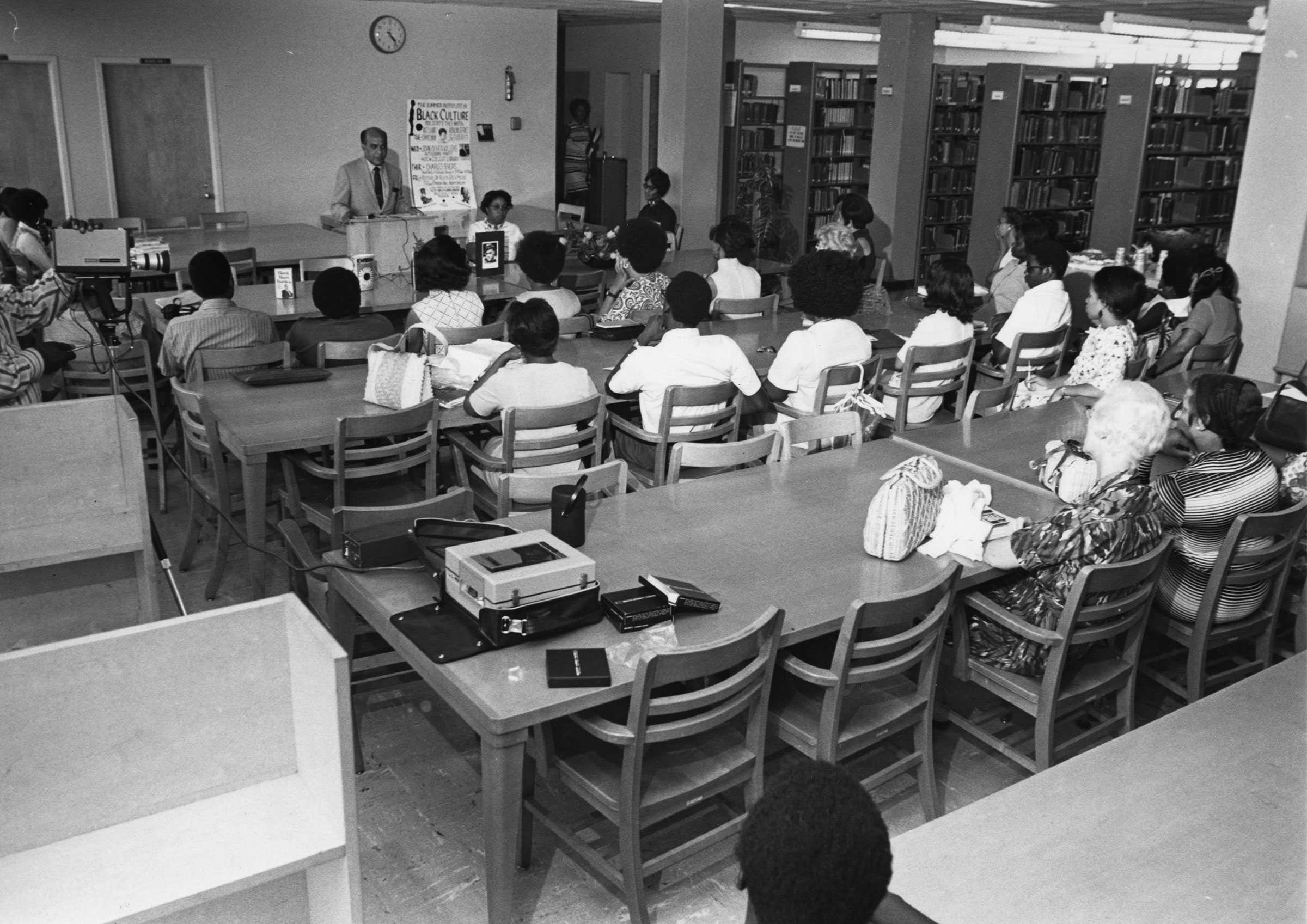 Photo of a group attending a lecture in a library, with a speaker at a podium and an audience seated at tables.