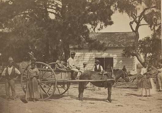 Photo shows a group of enslaved Black people posed around a horse-drawn cart