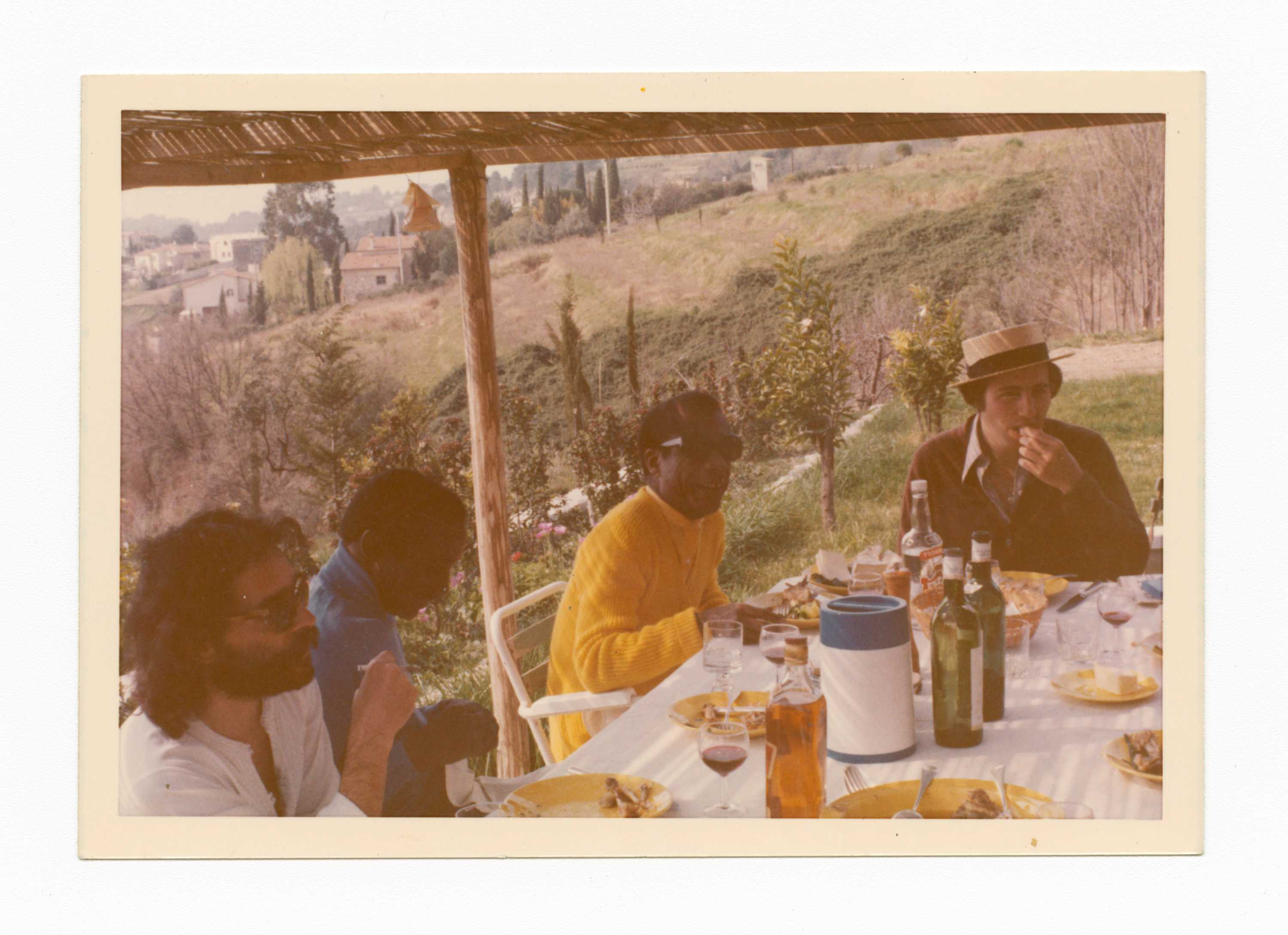 A colored, but faded photograph of James Baldwin and three friends sitting outside around a table.