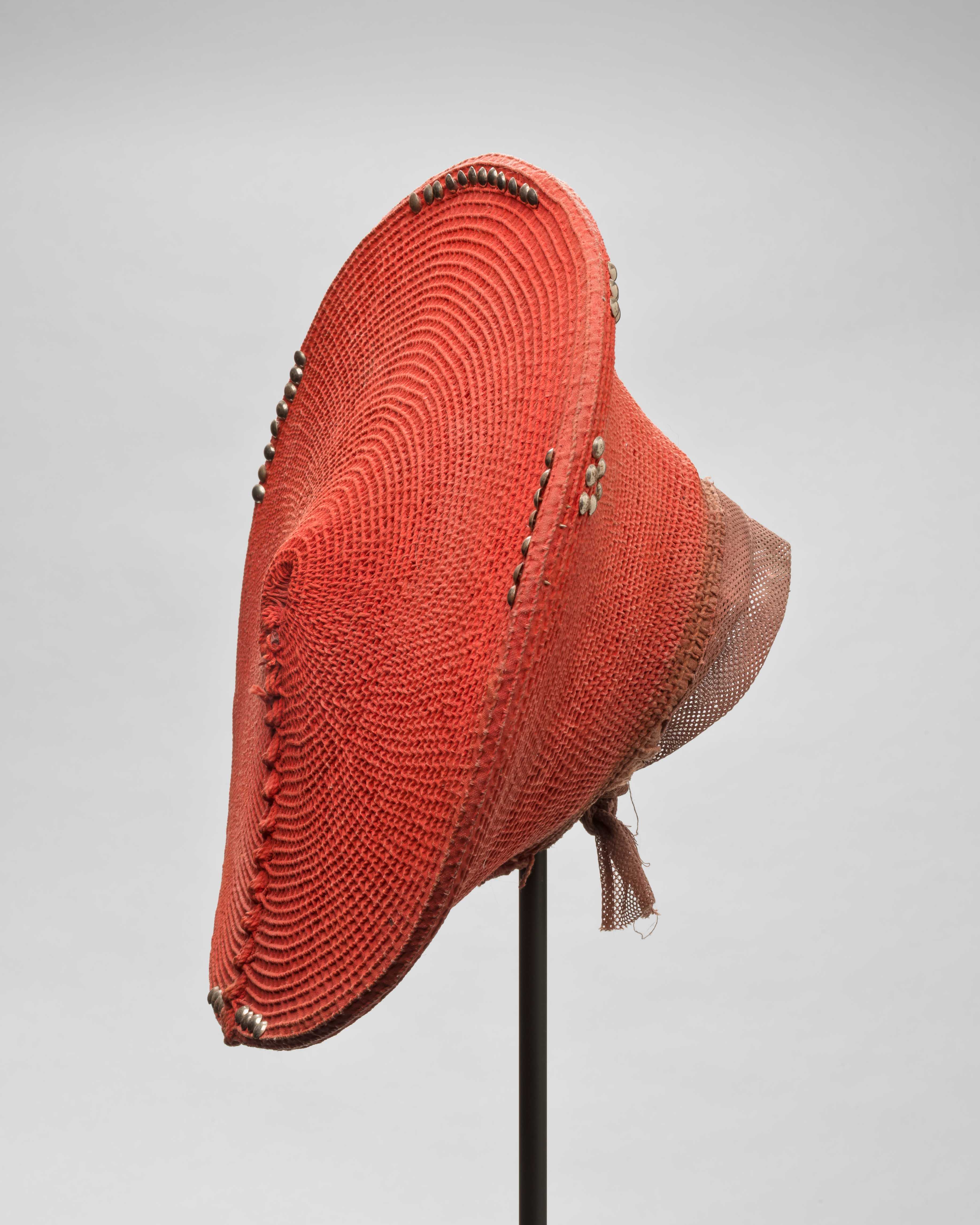 A woven, coral colored hat made of grass fiber, fat, ochre, metal, and pigment.