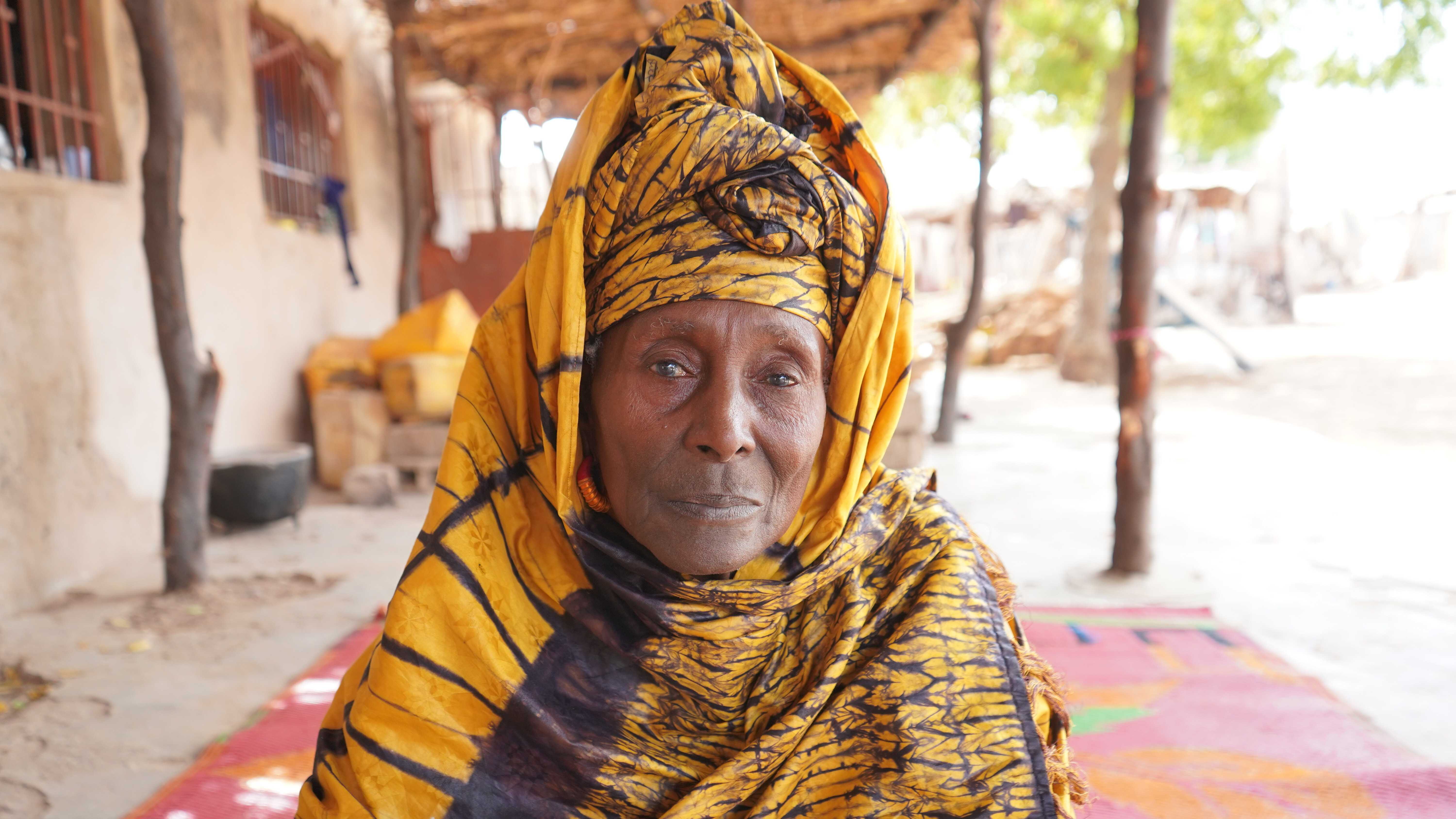Close-up portrait of a person wearing a bright yellow patterned headwrap and matching shawl, seated outdoors in a shaded courtyard with a blurred building and trees in the background.