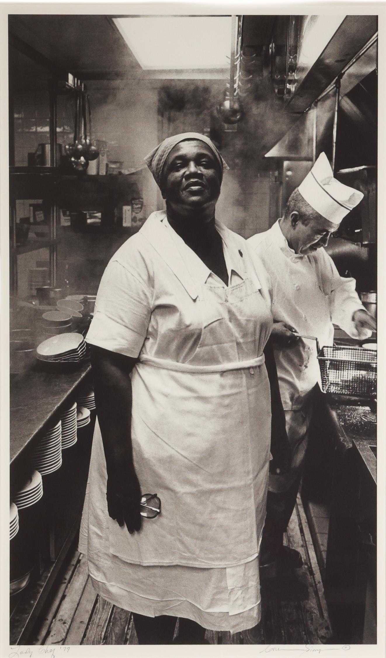 Black and white photograph of a female chef standing in her kitchen.  She is dressed all in white with a kerchief on her head.