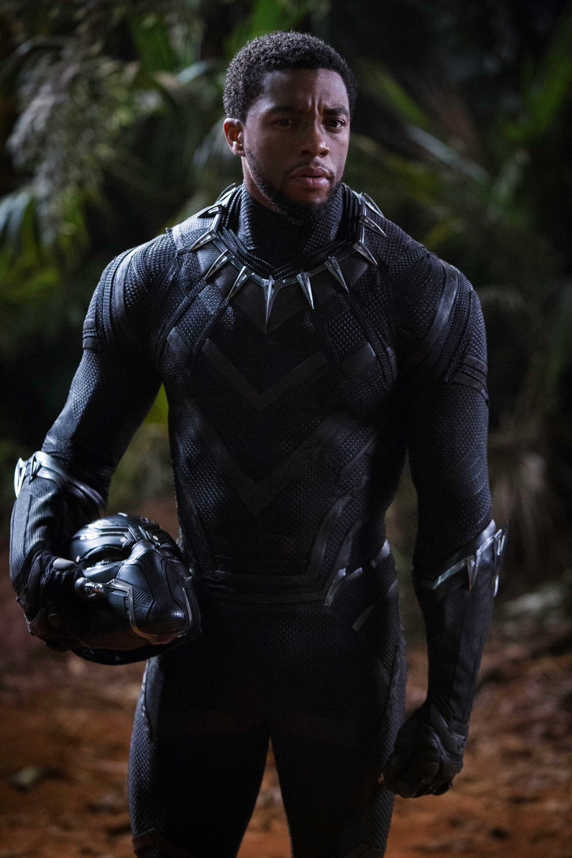 Chadwick Boseman, playing the Black Panther, stands in his suit in a forest, with his helmet off. He is resting it against his hip.