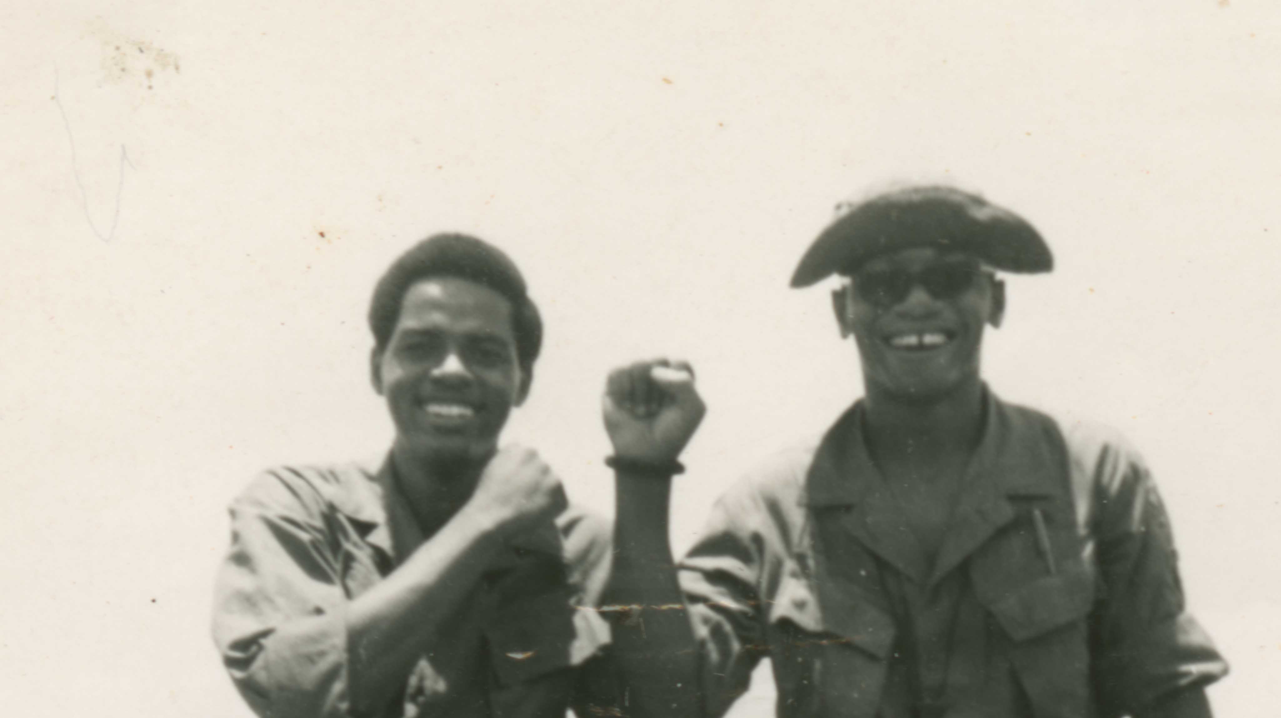 A black and white photograph of two soldiers, in military fatigues, perched on the edge of the windshield, boots resting on the jeep's hood.