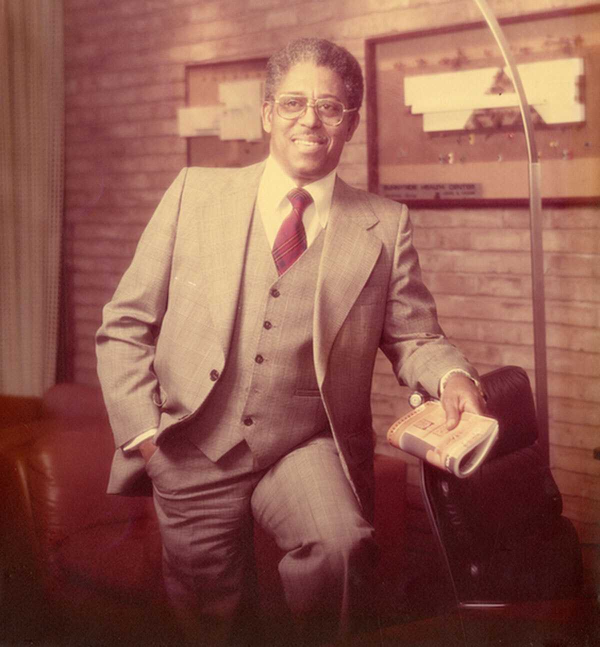 Architect John Chase wearing a three-piece suit and glasses, holding a rolled-up newspaper while leaning against a chair. The background includes brick walls with framed items and lighting fixtures, suggesting a formal or office setting.