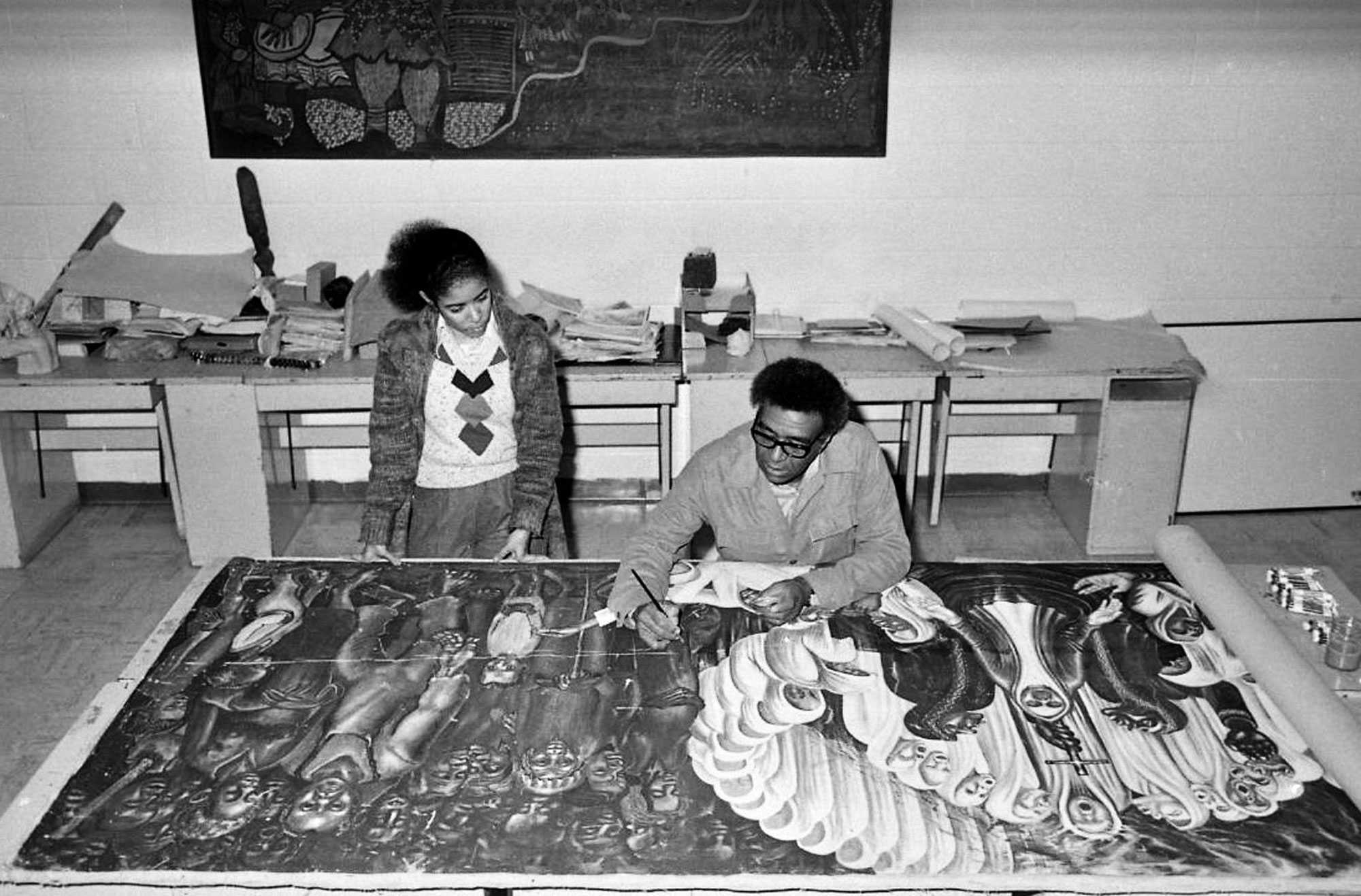 Black-and-white photograph of two people working on a large artwork spread across a table, surrounded by papers and art supplies.