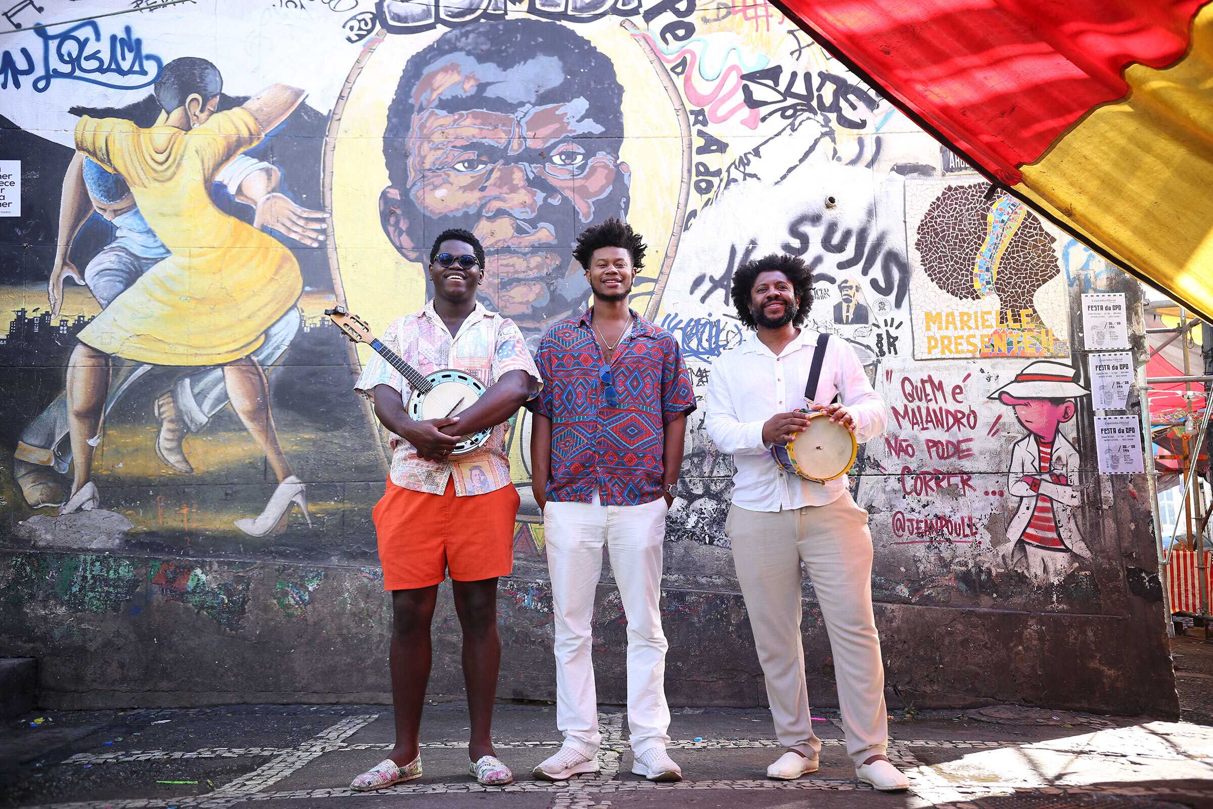 Alcino Amaral stand with two other men in front of a mural. The two men hold various instruments.