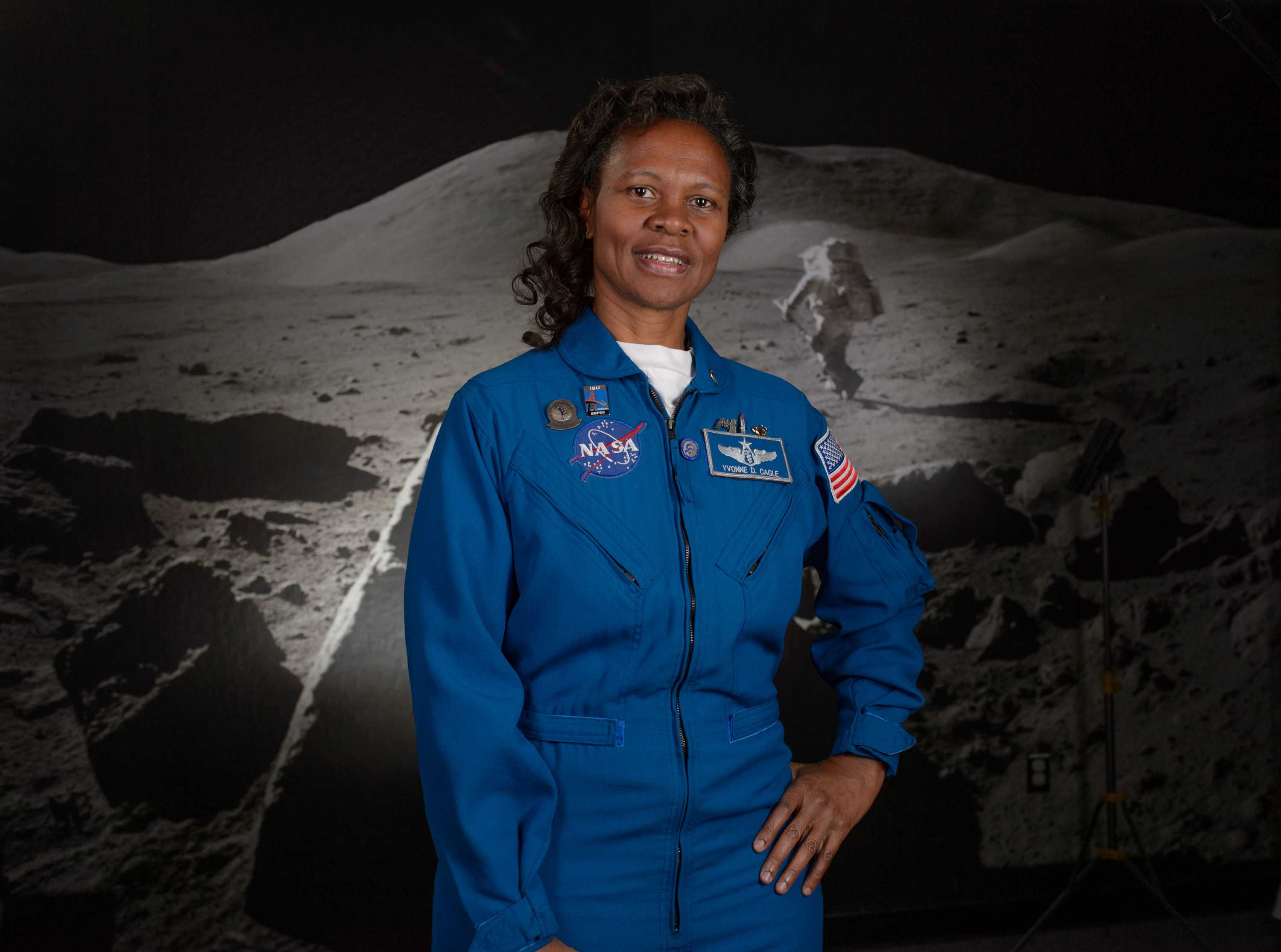 Yvonne Cagle poses with one hand on her hip in a blue NASA suit in front of a moon background.