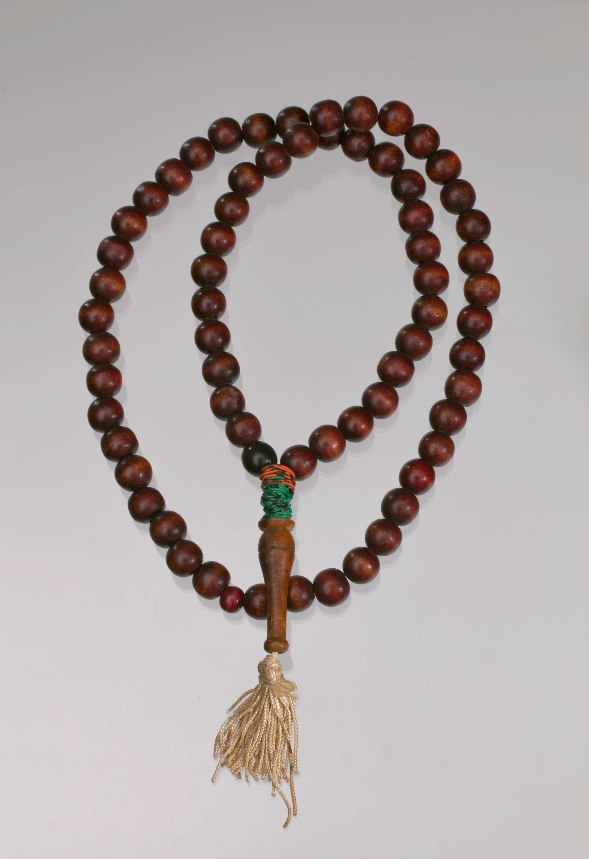 Brown beaded necklace made of 68 round wooden beads. Wood piece with tan tassel at end of necklace. Orange and green wire near tassel.