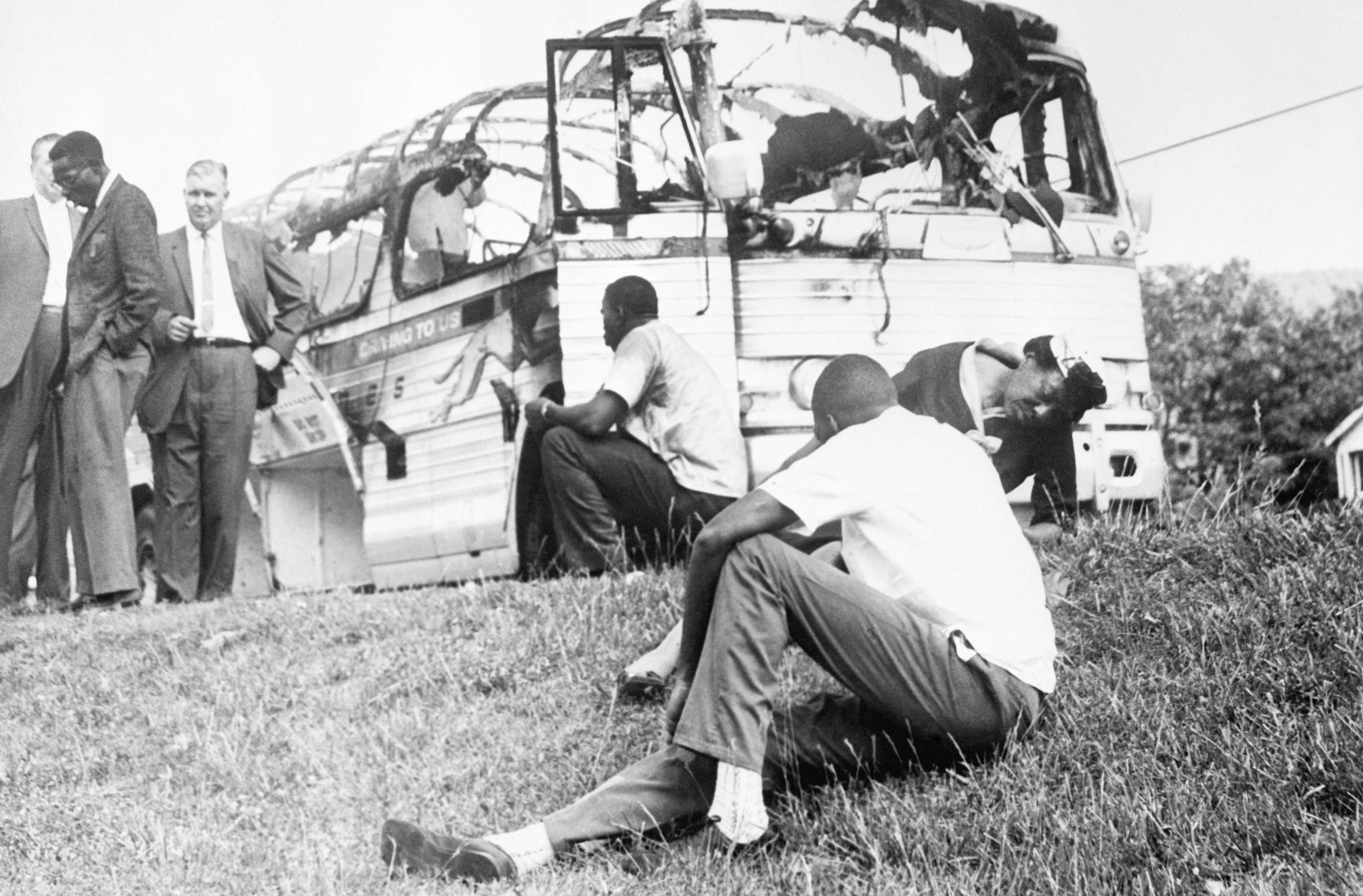 Black and White Photograh of men standing and sitting near burned out bus