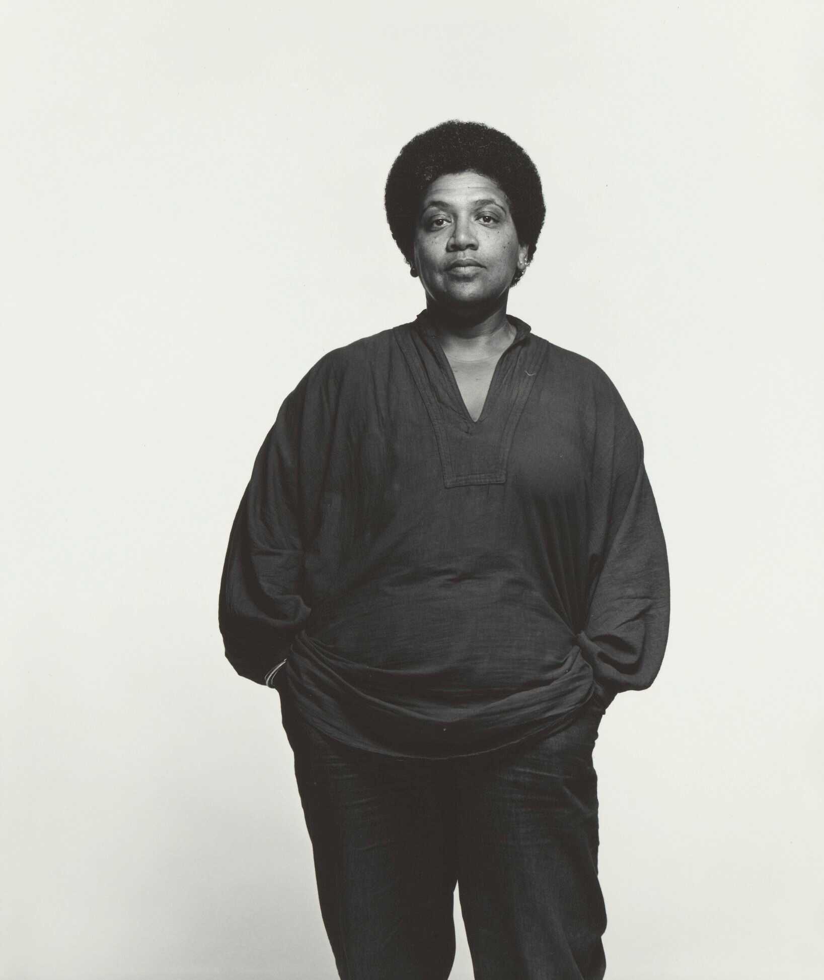 A black and white photograph of Audre Lorde. She is featured standing, facing the camera with her hands in her pockets. The back of the photograph has caption information at center and the number [3034] in the top left corner.