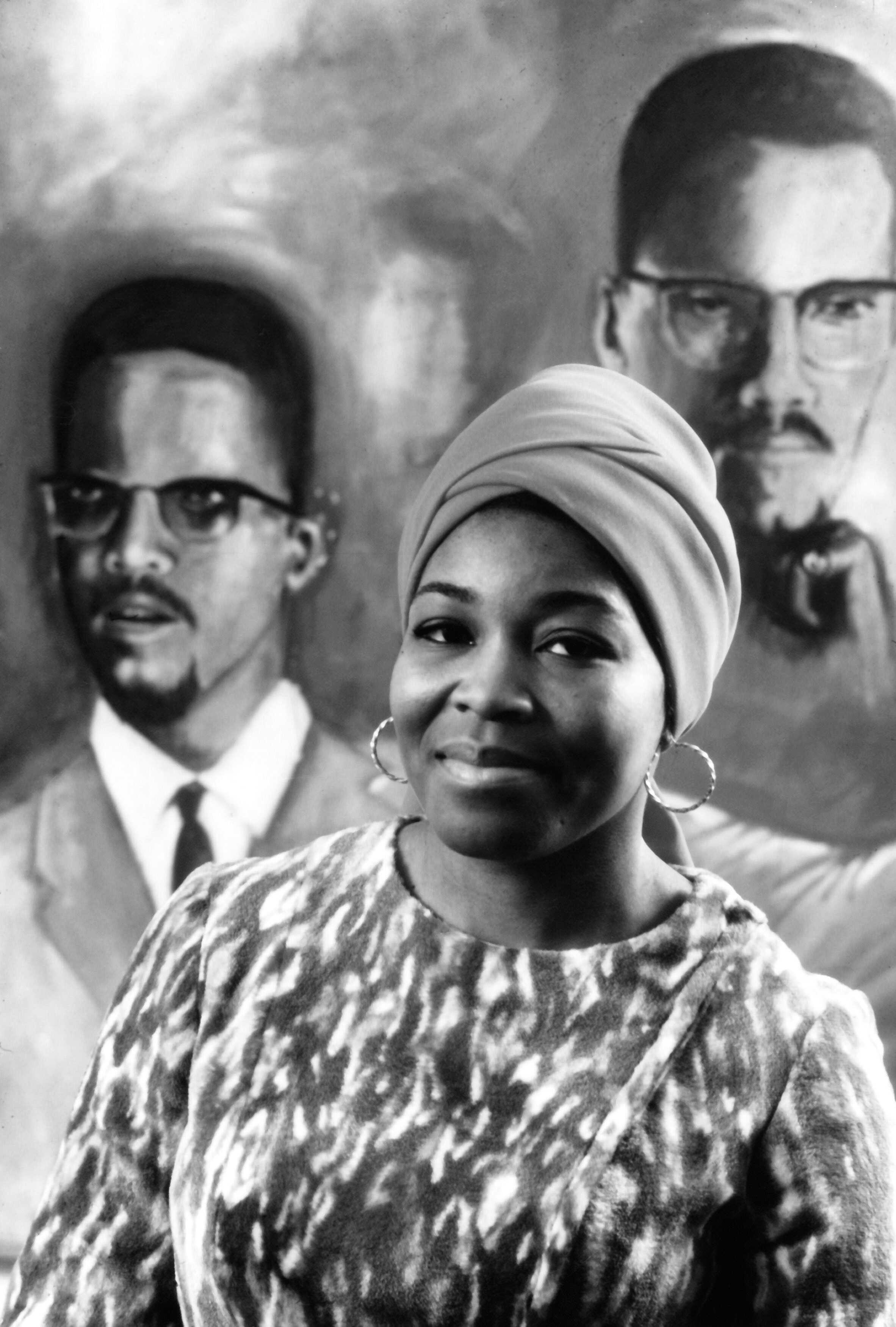 Black and white photograph of Betty Shabazz standing in front of a portrait of her late husband, Malcolm X