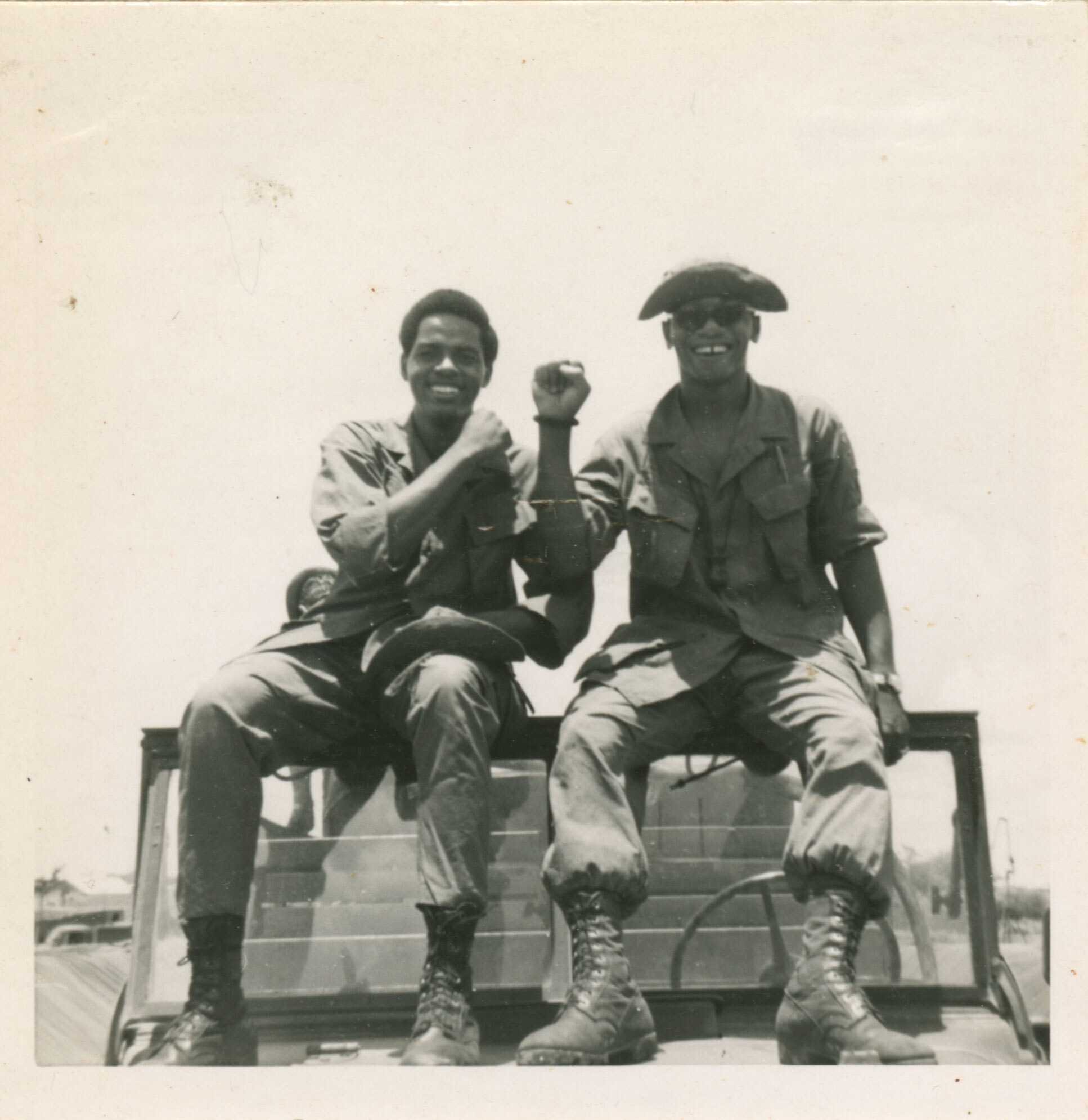 A black and white photograph of two soldiers, in military fatigues, perched on the edge of the windshield, boots resting on the jeep's hood.
