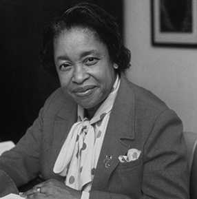 Author Margaret Walker seated at a desk wearing a suit jacket and polka dot scarf, looking at the camera with a slight smile.