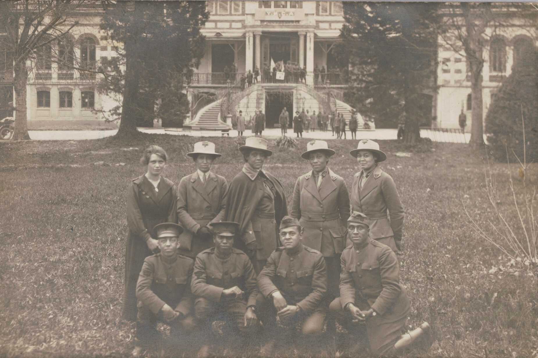 A real photo postcard of a group of military service men and women taken at the YMCA camp near Chambery, France, during World War I. The image depicts five women standing in a row on a lawn, with four men crouched in a row in front of them. Addie Waites Hunton is in the center of the back row; the other women and men are unidentified. In the background is a large building with a double staircased entrance. A temporary sign reading [Y.M.C.A.] has been placed on the portico at the top of the stairs. Other individuals are visible along the top and bottom of the stairs. The verso has printing reading [CARTE POSTALE] with spaces for [Correspondance] and [Adresse] and a horse and horsehead mark for the publisher Guilleminot. The postcard has not been sent, but there is an inscription across the back by hand in brown ink reading [From Sgt. Thomas, who / was on leave at colored, / Y.M.C.A. at Chamberry / France]. There is an inscription by a different hand in graphite above the [Adresse] label reading [(ALFRED JACK THOMAS)].